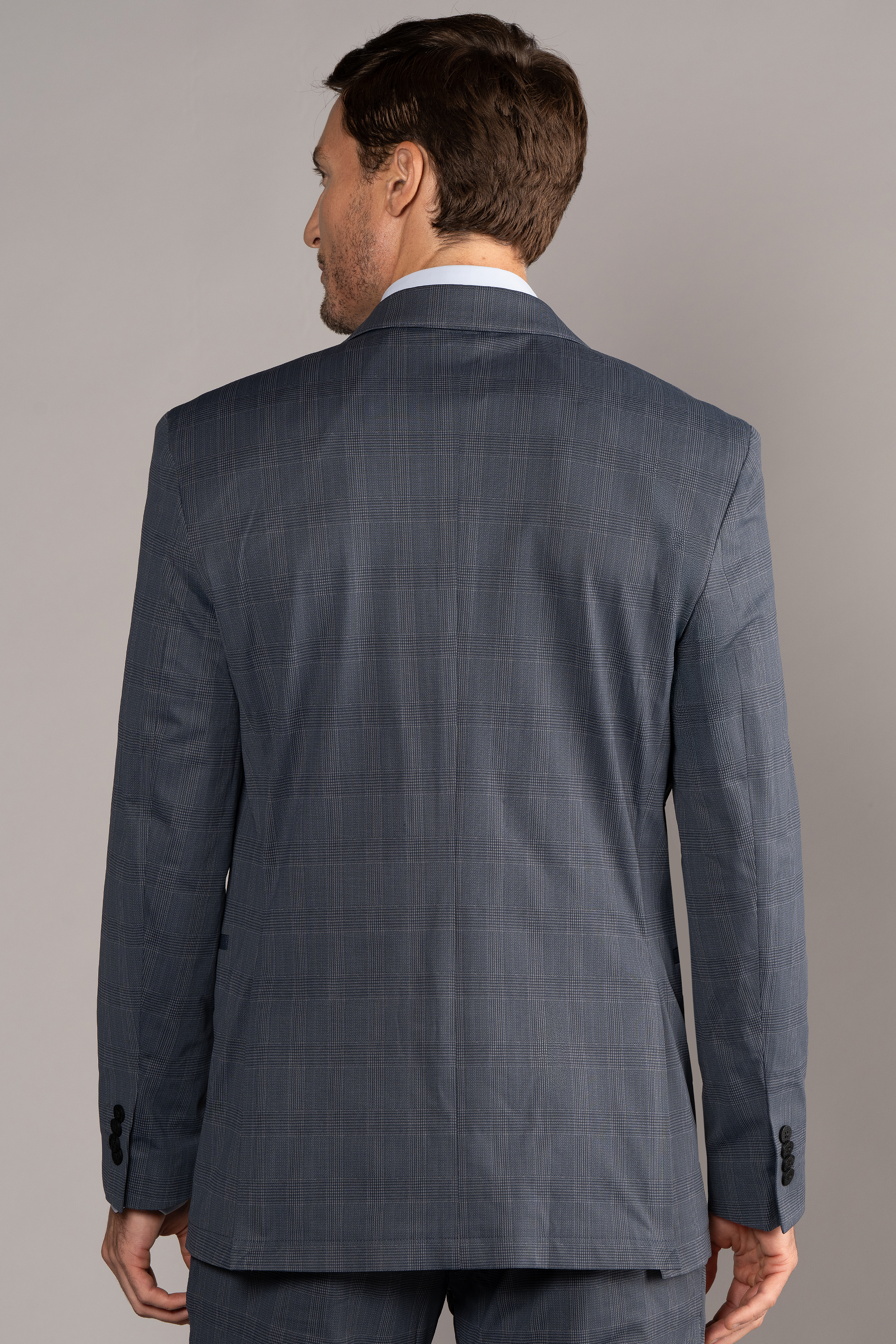 A man faces away, displaying the back of Twillory's Performance Blazer // Grey Prince of Wales. The stretch blazer features 3D shoulder pads, buttoned cuffs, and a structured fit. He stands relaxed against a plain, light background.
