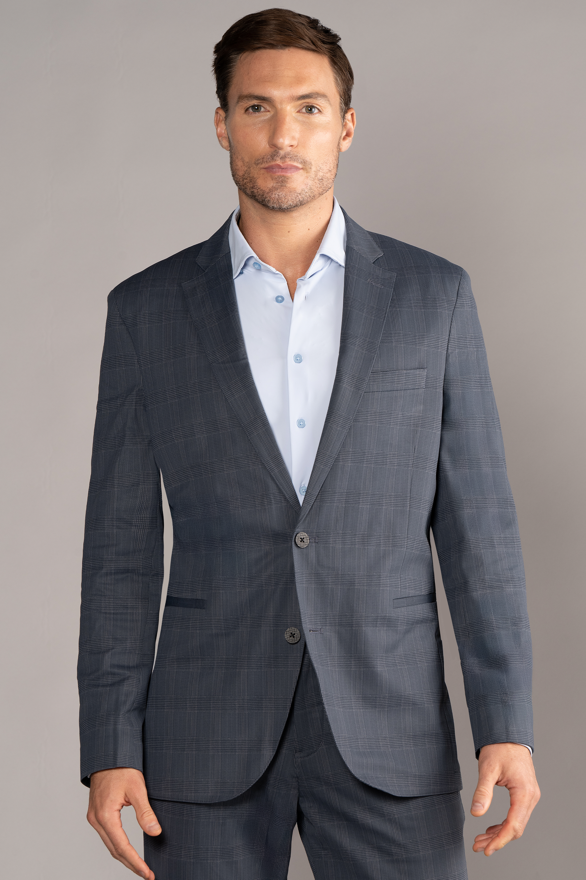 A man with short brown hair, light skin, and a trimmed beard faces forward in a Twillory Performance Blazer // Grey Prince of Wales over a light blue dress shirt. A plain background highlights his polished, professional style.
