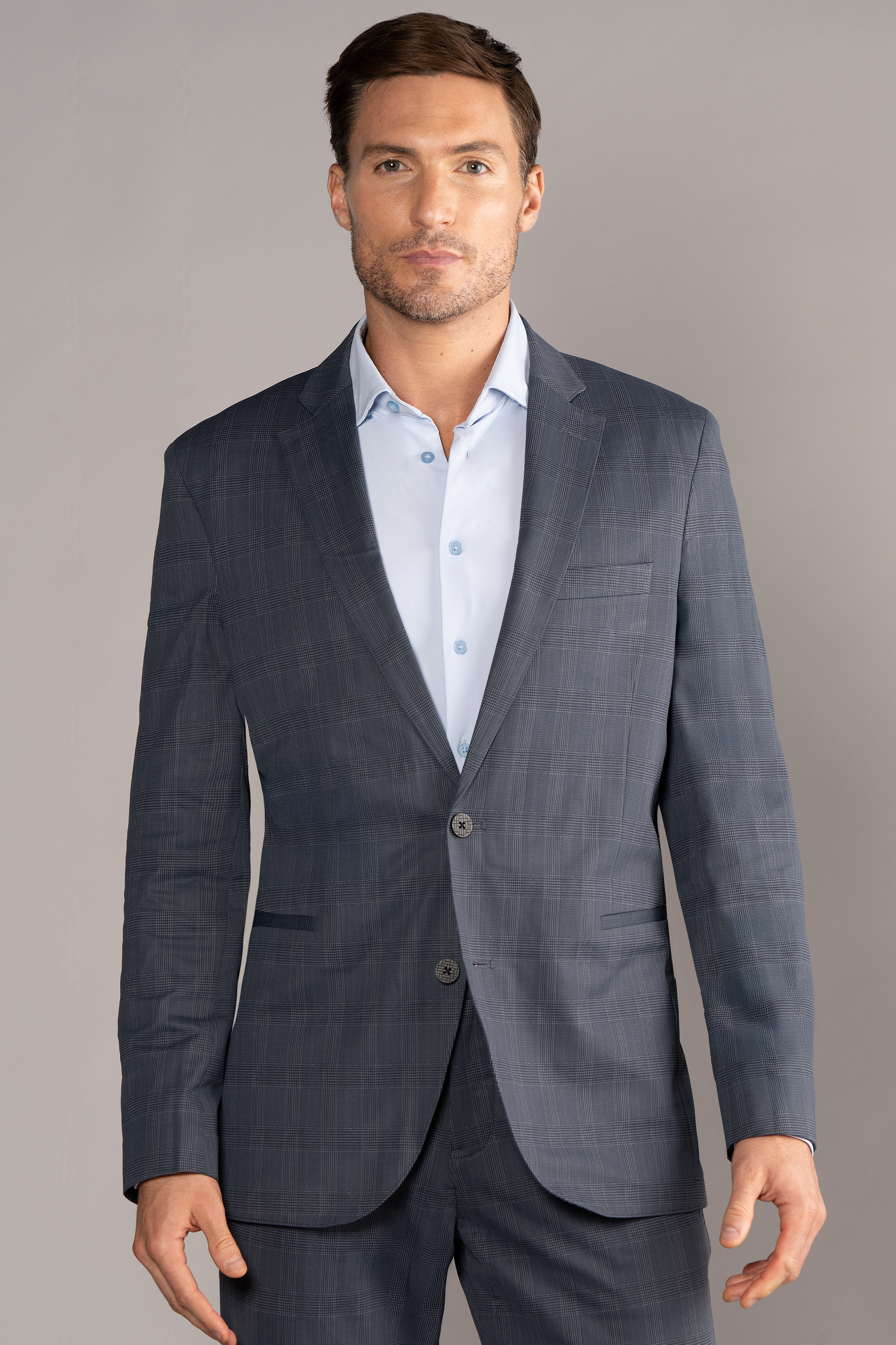 A man with short brown hair, light skin, and a trimmed beard faces forward in a Twillory Performance Blazer // Grey Prince of Wales over a light blue dress shirt. A plain background highlights his polished, professional style.