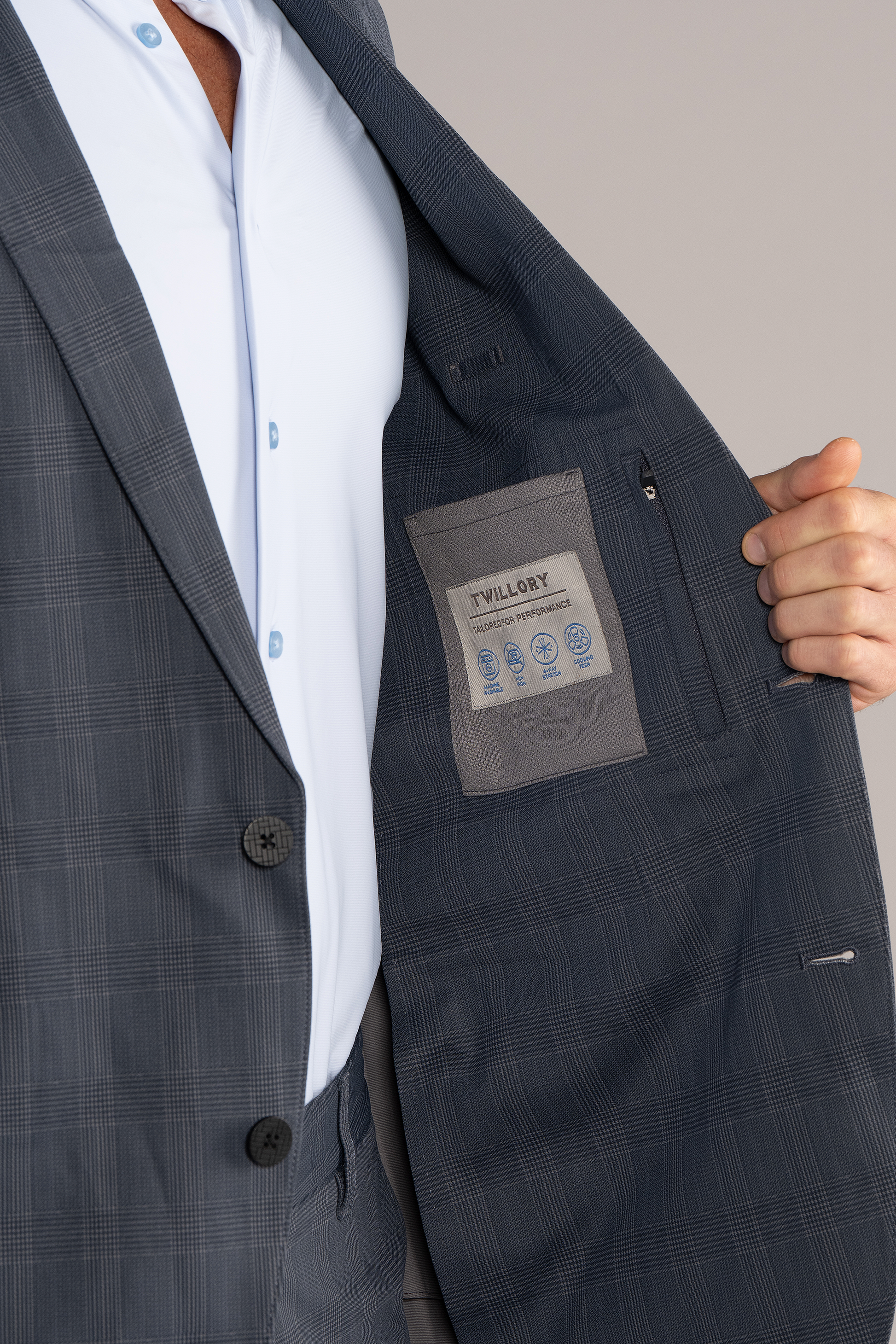 A person wearing the Twillory Performance Blazer // Grey Prince of Wales holds it open to show the inner pocket and a Twillory label sewn onto gray fabric. The light blue shirt underneath is also partially visible.