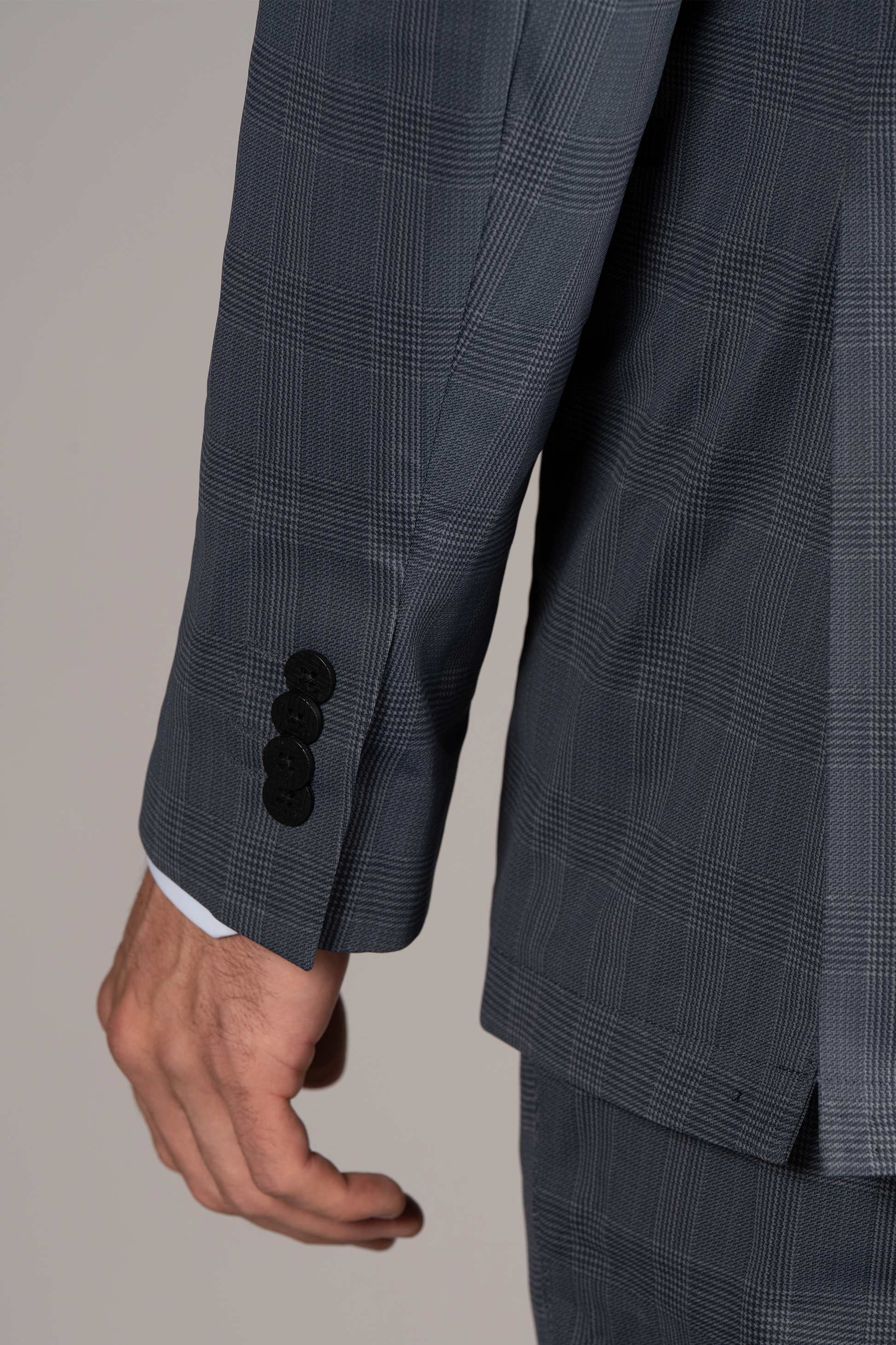 A close-up of a person's left arm in the Twillory Performance Blazer // Grey Prince of Wales, worn over a white shirt. Four black buttons accent the sleeve near the wrist, with relaxed fingers against a plain light background.