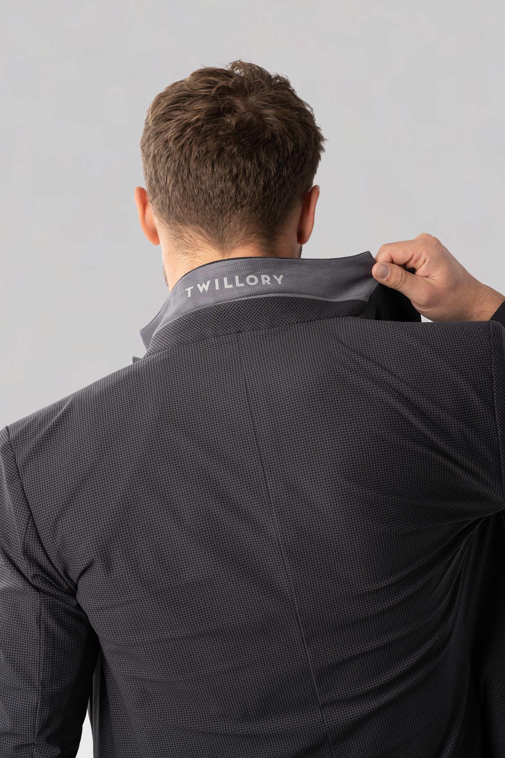 A person, seen from behind, lifts the collar of a dark grey houndstooth Twillory Performance Blazer, revealing "TWILLORY" printed in white on the inside. The background is plain and light gray.