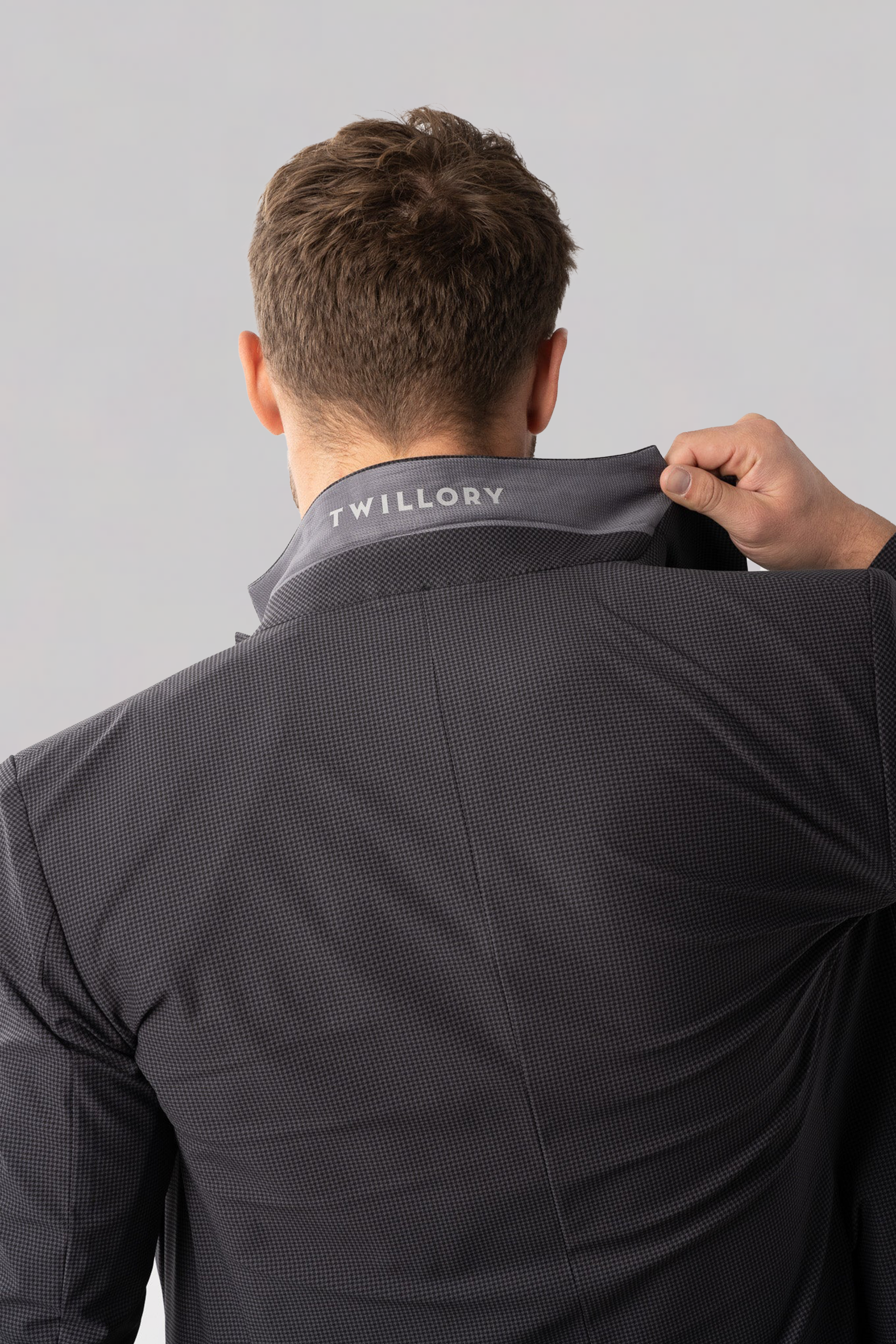 A person, seen from behind, lifts the collar of a dark grey houndstooth Twillory Performance Blazer, revealing "TWILLORY" printed in white on the inside. The background is plain and light gray.