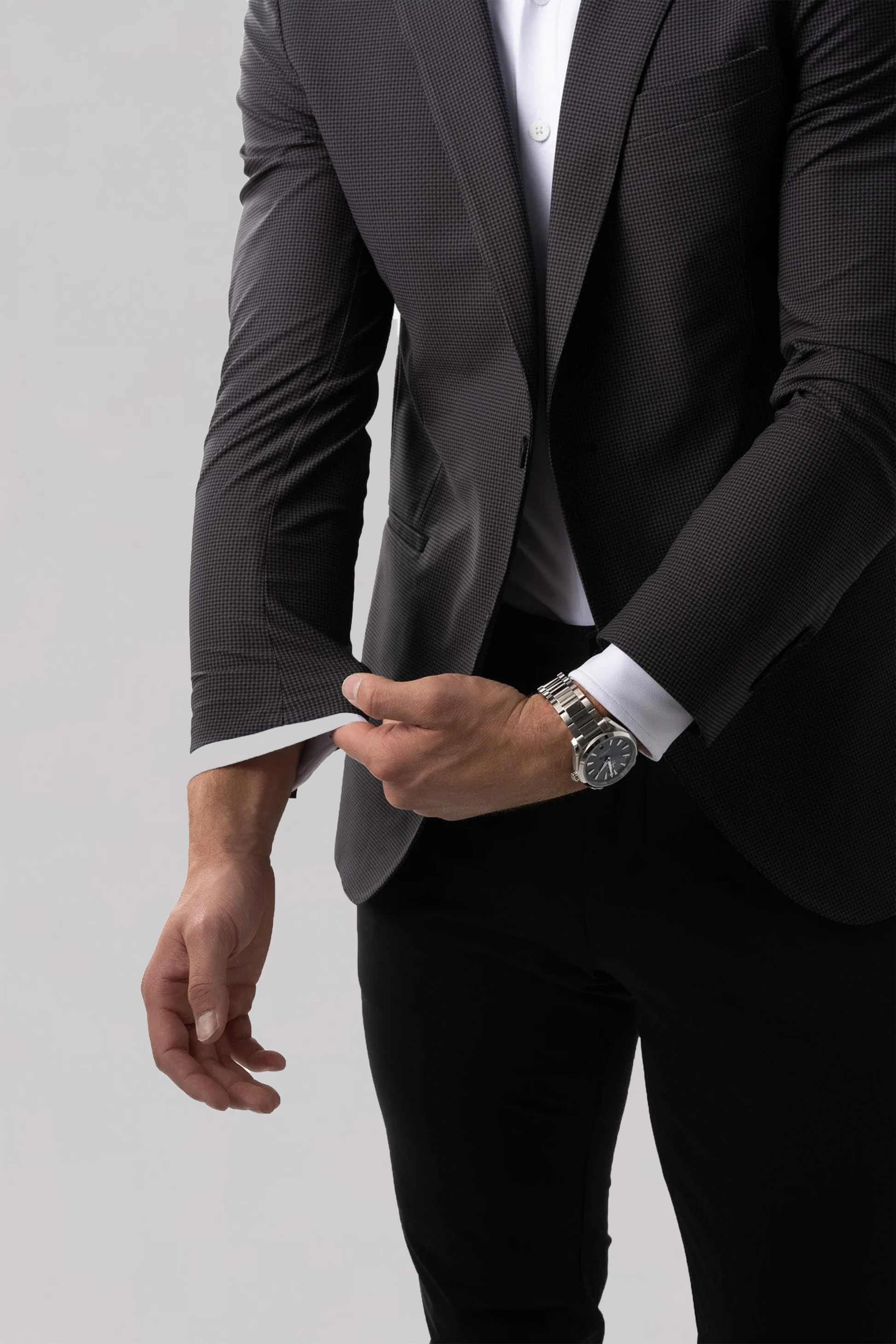 A man in a Twillory Performance Blazer in Grey Houndstooth adjusts his sleeve over a white dress shirt and black pants. He wears a silver watch on his left wrist against a plain light gray background, with his head out of frame.