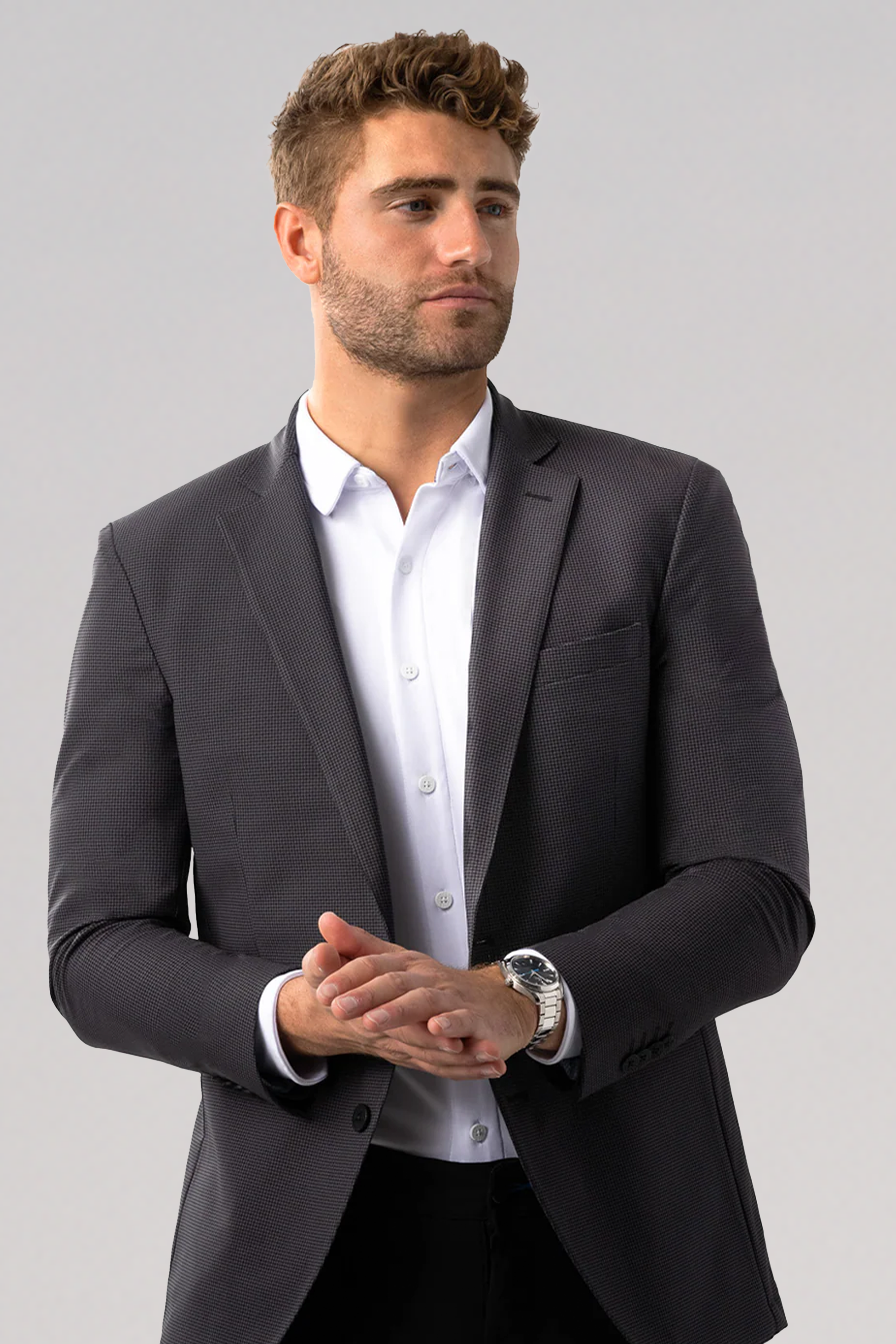 A man with short, wavy brown hair and a trimmed beard wears the Twillory Performance Blazer // Grey Houndstooth over a white dress shirt. He stands against a plain light gray backdrop, looking slightly to the side with hands clasped at his waist.