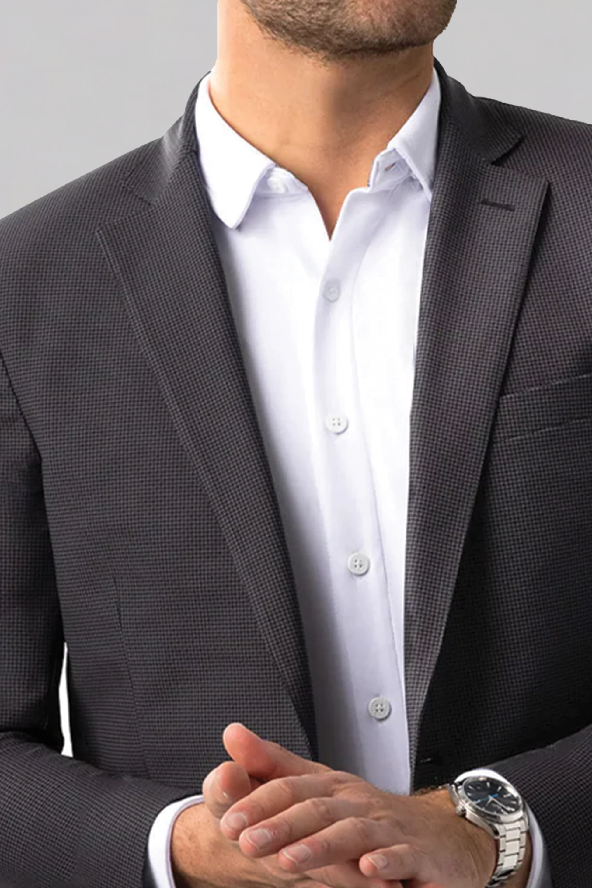 A man wears the Twillory Performance Blazer // Grey Houndstooth over a crisp white dress shirt. His clasped hands show a silver wristwatch. The photo is cropped at the nose and mid-torso against a plain light gray background.