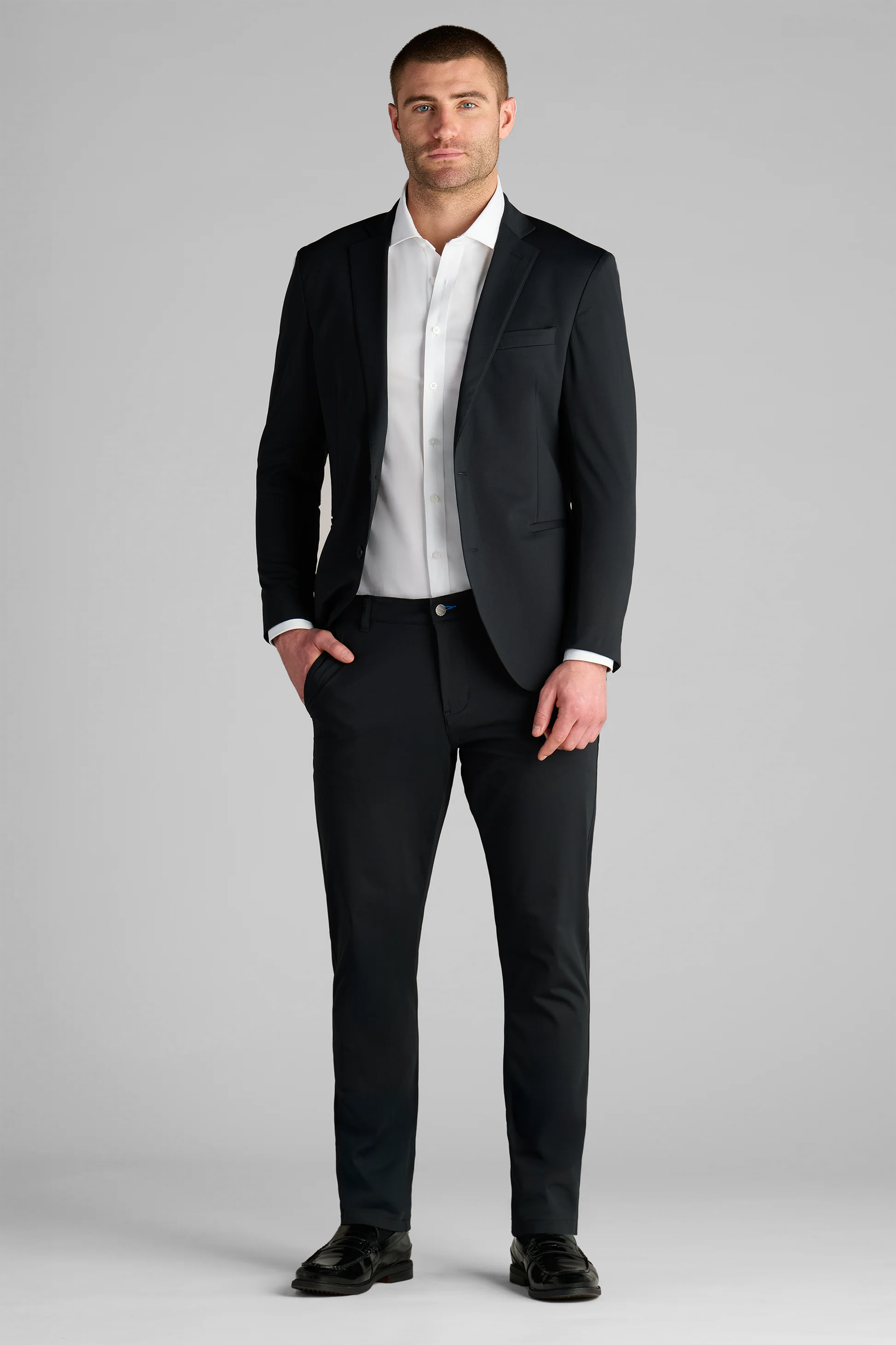 A man with short hair and a trimmed beard stands facing forward against a gray background, wearing the Performance Blazer // Black—a fitted, wrinkle-free blazer with performance stretch—over a white shirt and black dress shoes. His left hand is in his pocket.