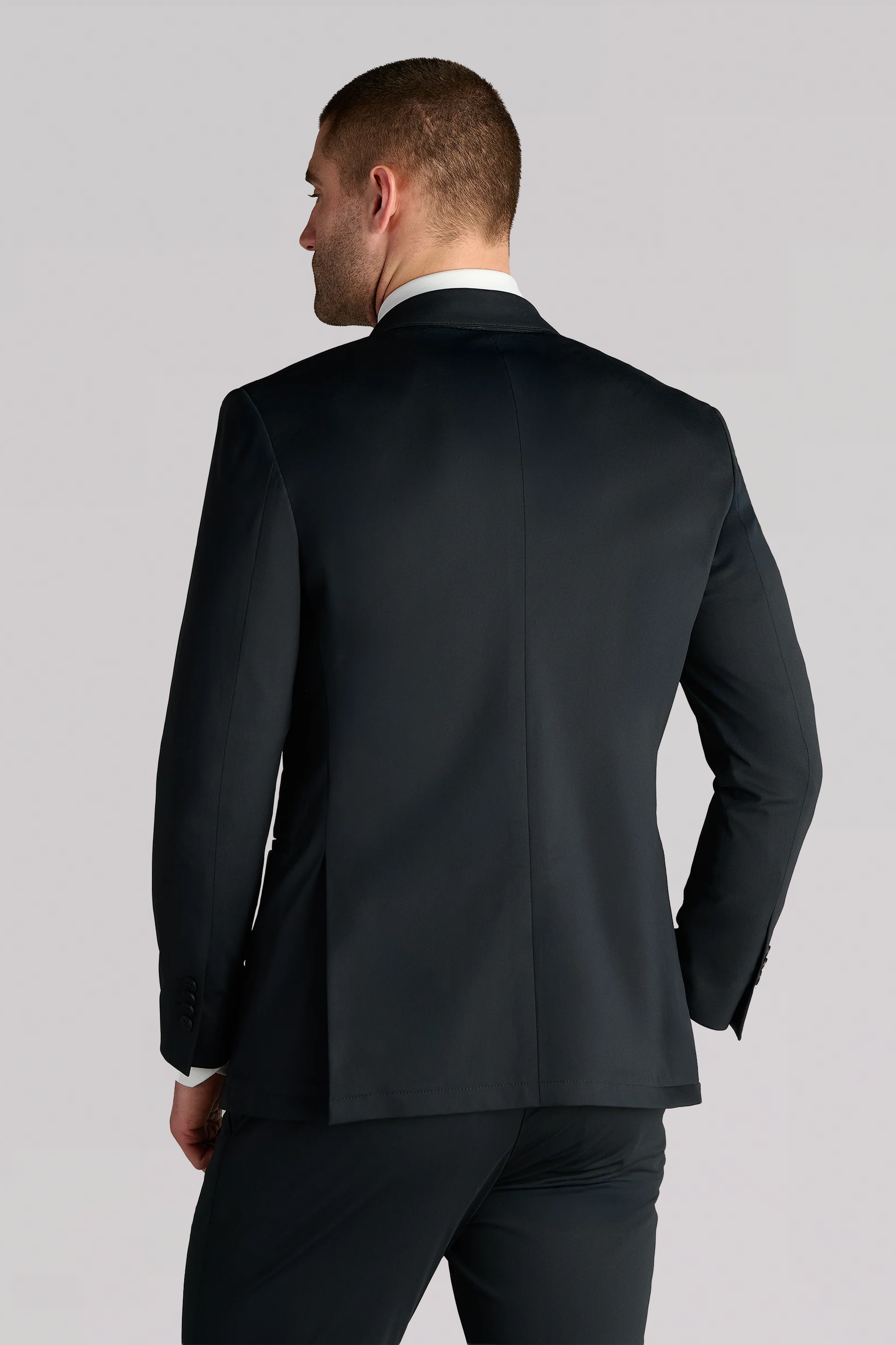 A man is seen from behind wearing the Performance Blazer // Black with matching trousers, standing against a plain light gray background.