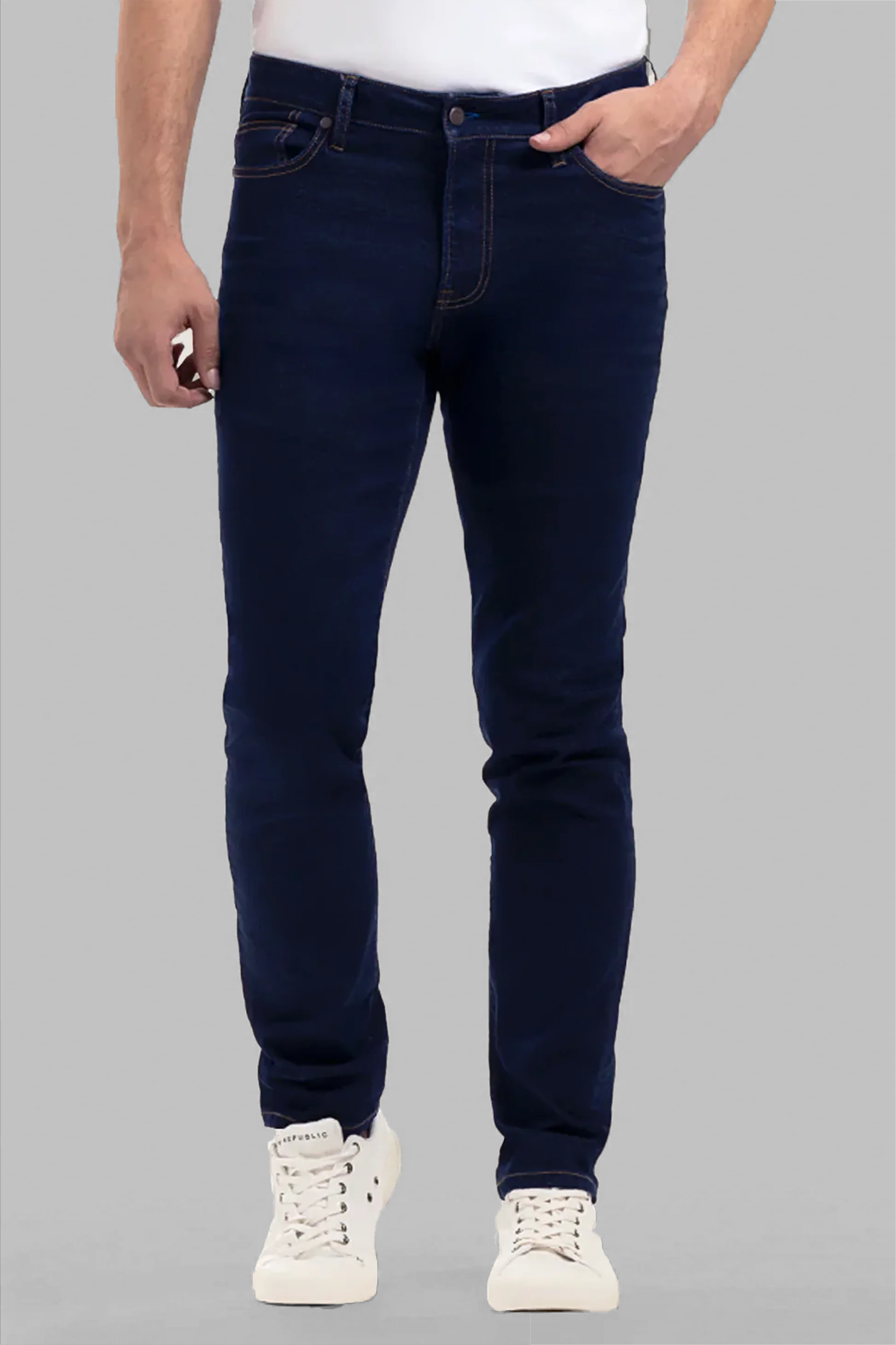 A person in Performance Denim // DARK WASH INDIGO slim-fit jeans, a white shirt, and white sneakers stands with one hand in their pocket against a plain grey background.
