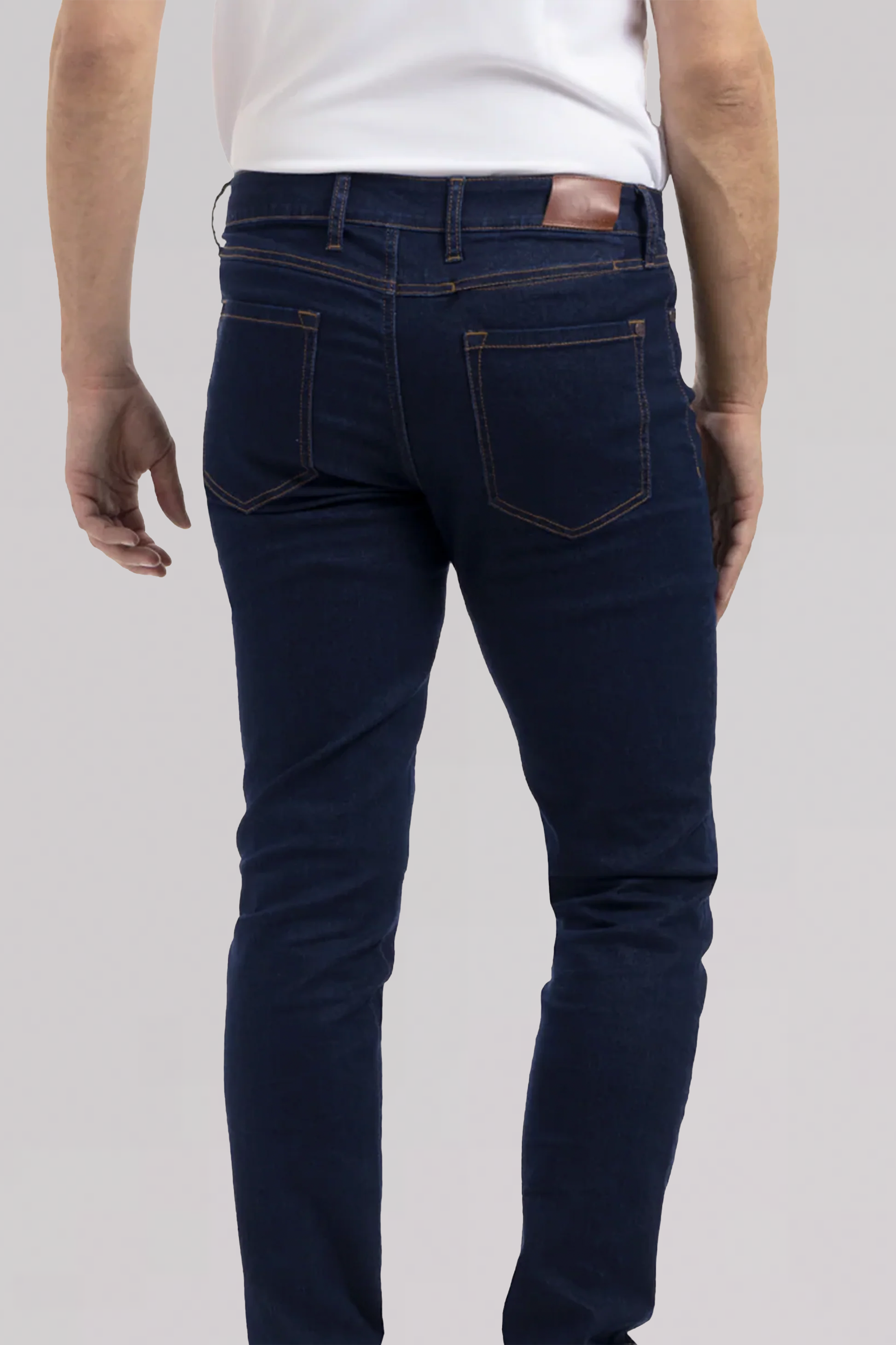 A person is shown from the back wearing Performance Denim // DARK WASH INDIGO jeans with two back pockets and brown stitching, paired with a white shirt. Their left arm is slightly bent while the right arm is relaxed.