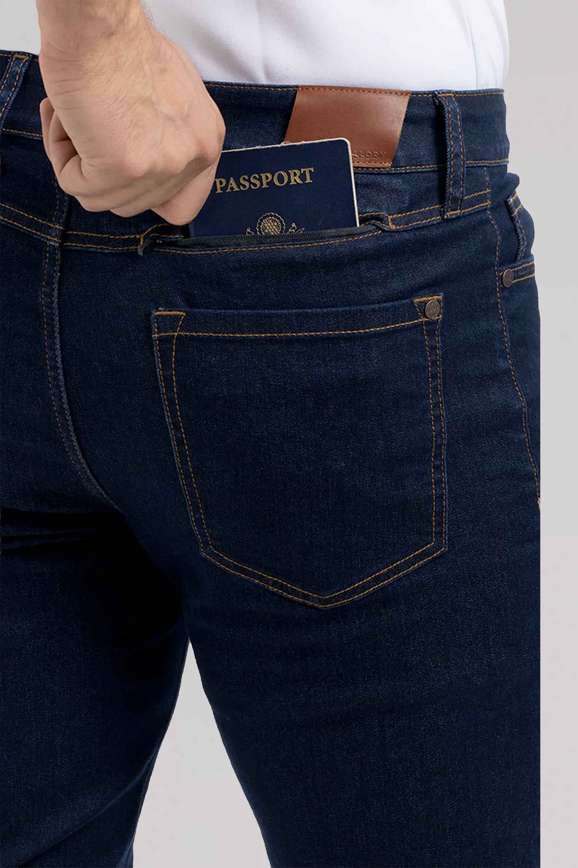 A person slips a dark blue passport into the back pocket of their Performance Denim // DARK WASH INDIGO jeans, showing only their lower torso and hand.