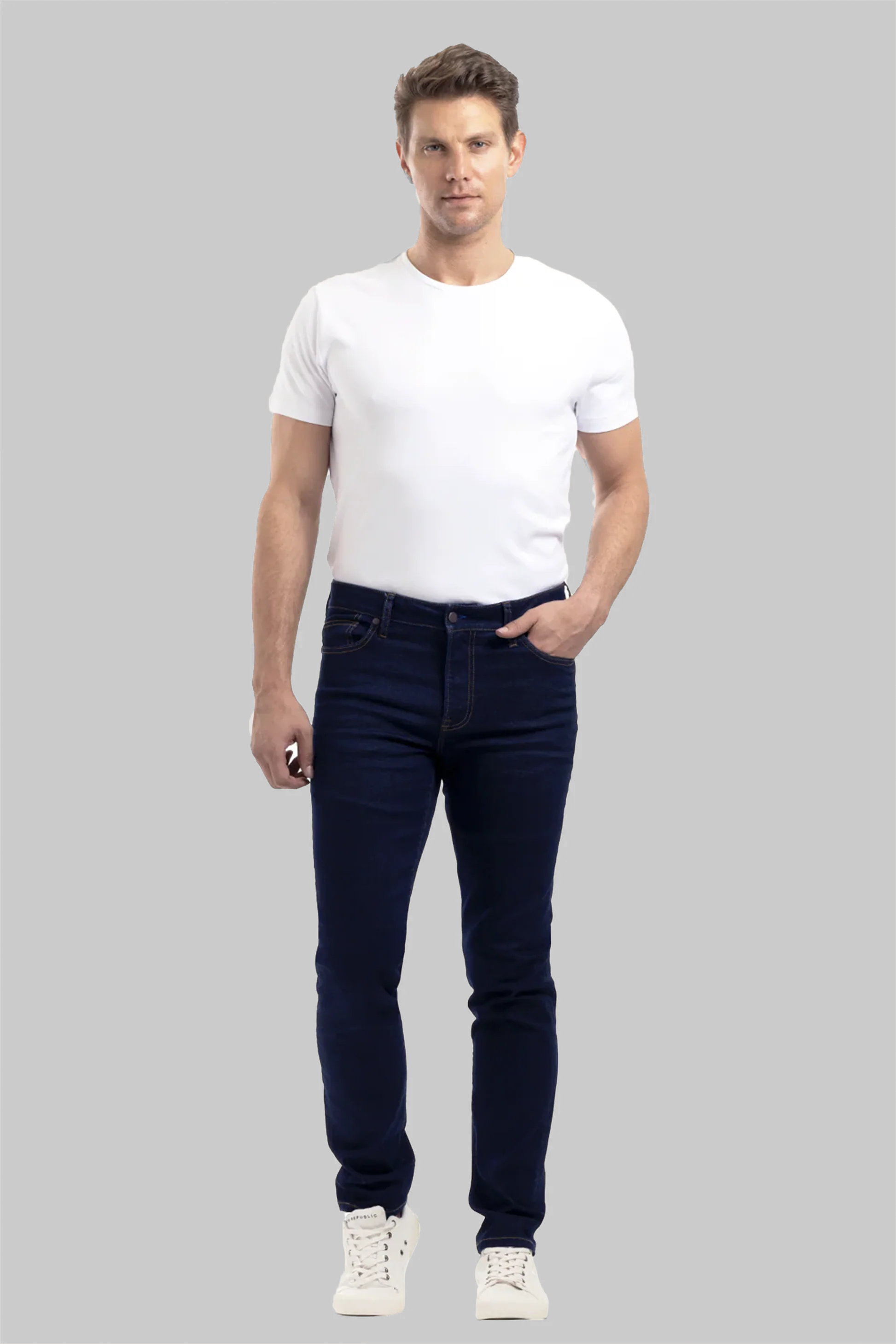 A man with short brown hair and a neutral expression stands facing forward against a grey background, wearing Twillory Performance Denim // DARK WASH INDIGO jeans, a fitted white t-shirt, and white sneakers. One hand is in his pocket.