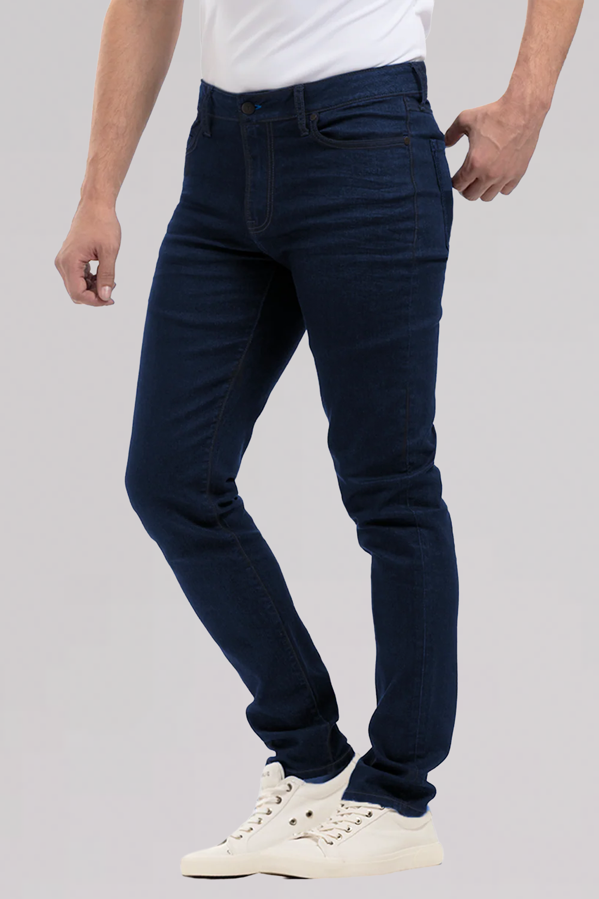 Against a light gray background, a person models Twillory’s Performance Denim // MIDNIGHT INDIGO jeans—tapered fit, dark blue—with a white T-shirt and sneakers. Only the lower torso, legs, and part of the arms are shown in a casual pose.