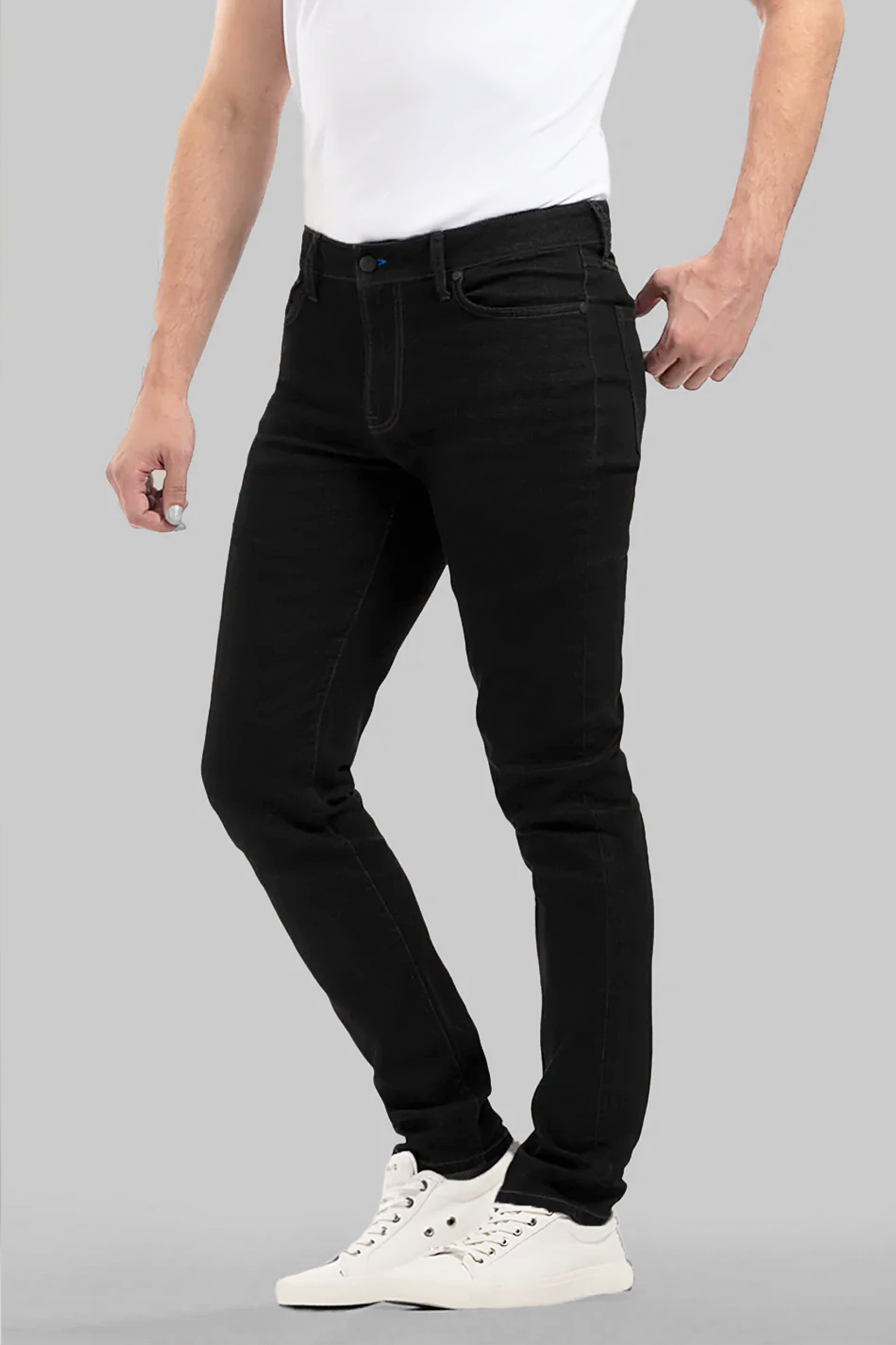 A person wears a fitted white t-shirt, Twillory Performance Denim // PURE BLACK jeans with a tapered fit, and white high-top sneakers against a plain gray background. Only the body from shoulders down is visible, one hand in a pocket.