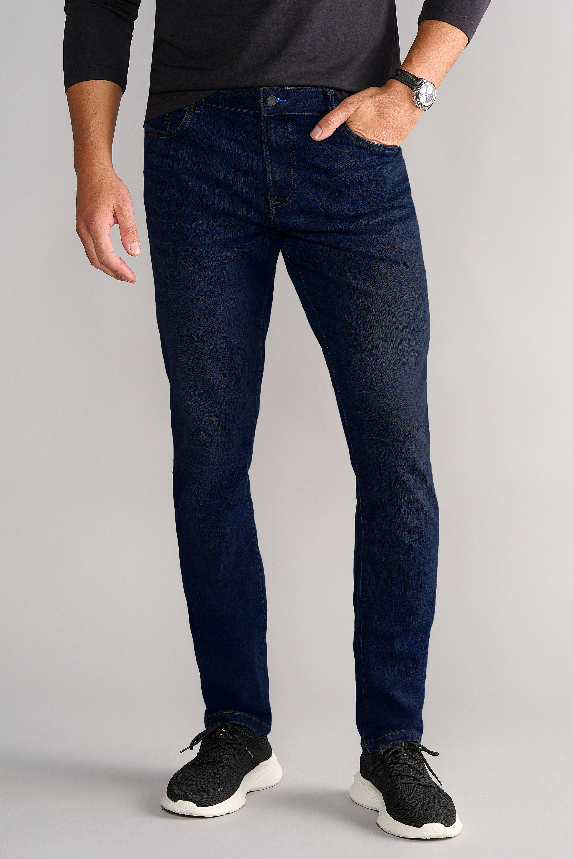A man stands against a light gray background in a long-sleeve black shirt, Performance Denim // MIDNIGHT INDIGO jeans with a Tapered Fit, black sneakers with white soles, and a wristwatch. His face is not visible.