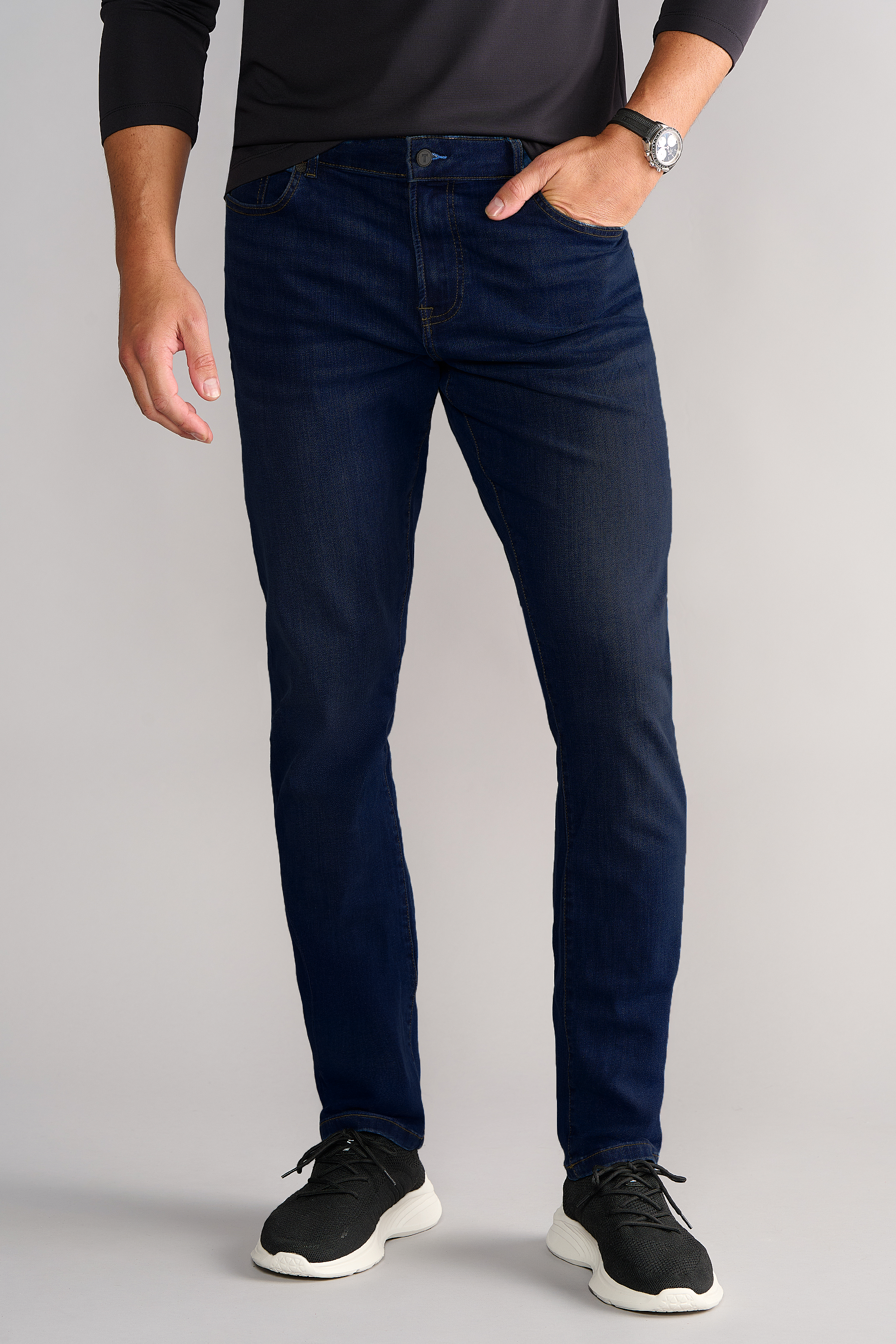 A man stands against a light gray background in a long-sleeve black shirt, Performance Denim // MIDNIGHT INDIGO jeans with a Tapered Fit, black sneakers with white soles, and a wristwatch. His face is not visible.