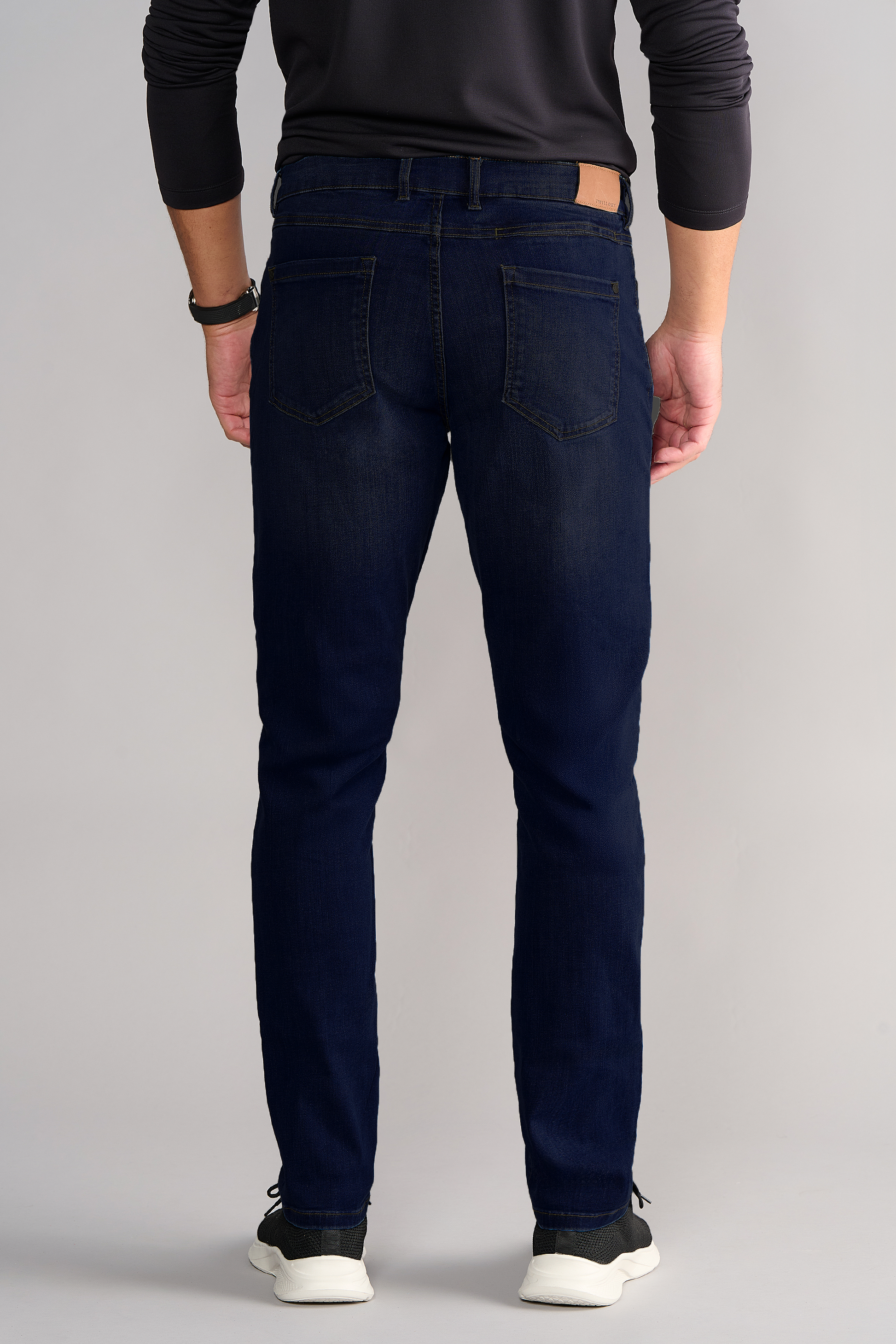 A person wearing Performance Denim // MIDNIGHT INDIGO with a tapered fit and a long-sleeved black top stands facing away, showing the two back pockets of the dark blue jeans paired with black sneakers with white soles.