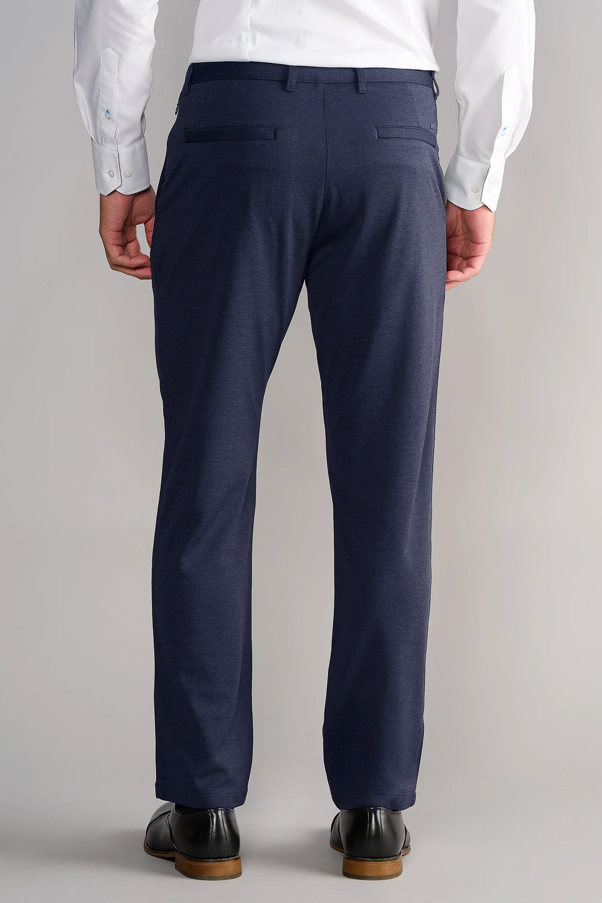 A person is seen from the back, wearing a white shirt tucked into Twillory Circular Knit Pants in Heather Navy. The wrinkle-resistant pants offer 4-way stretch, welt pockets, and are styled with black dress shoes with brown soles on a light gray background.
