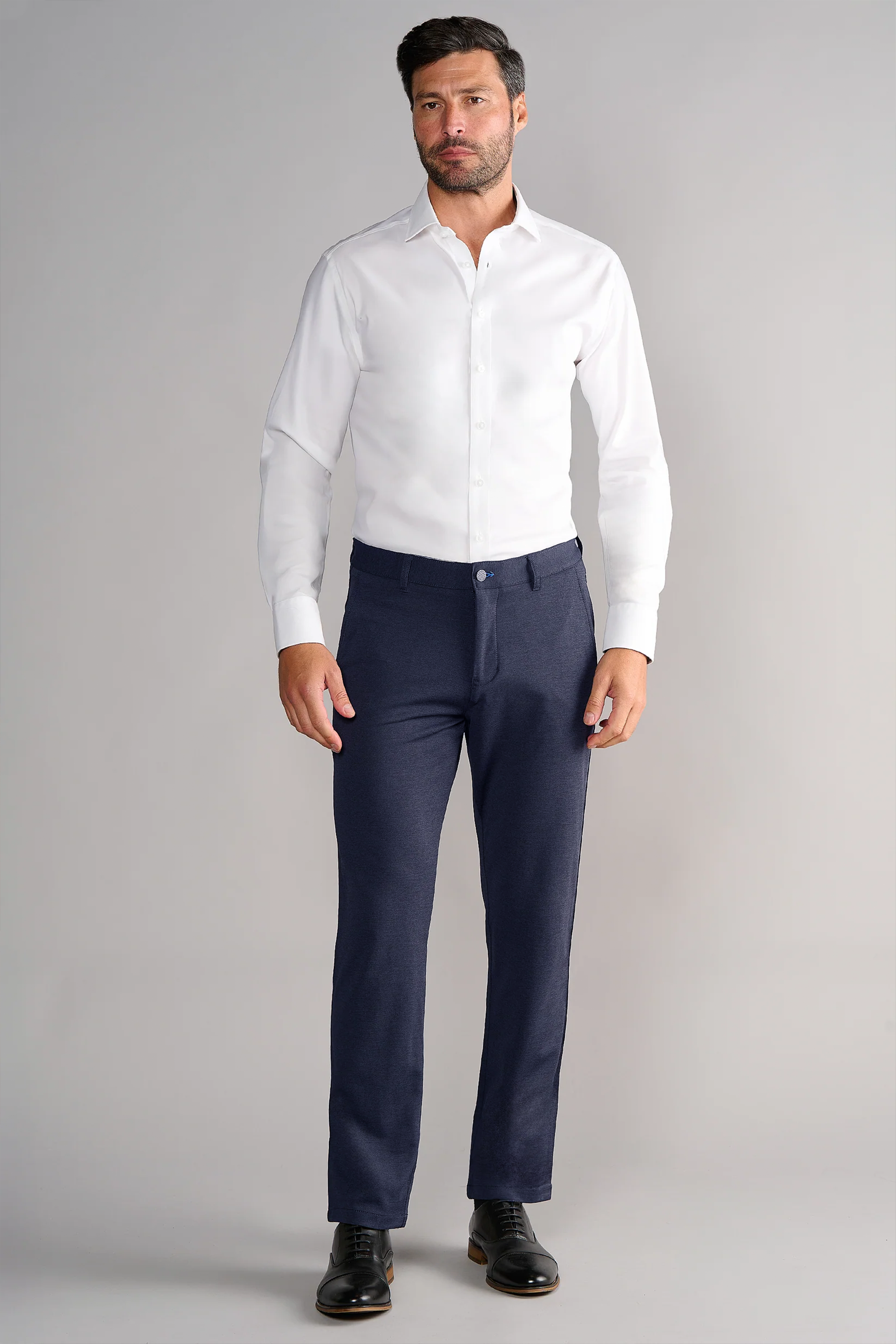 A man stands facing forward against a plain gray background. He wears a white button-up shirt and Twillory Circular Knit Pants in Heather Navy, known for their wrinkle resistance and 4-way stretch comfort. His expression is neutral, arms at his sides.