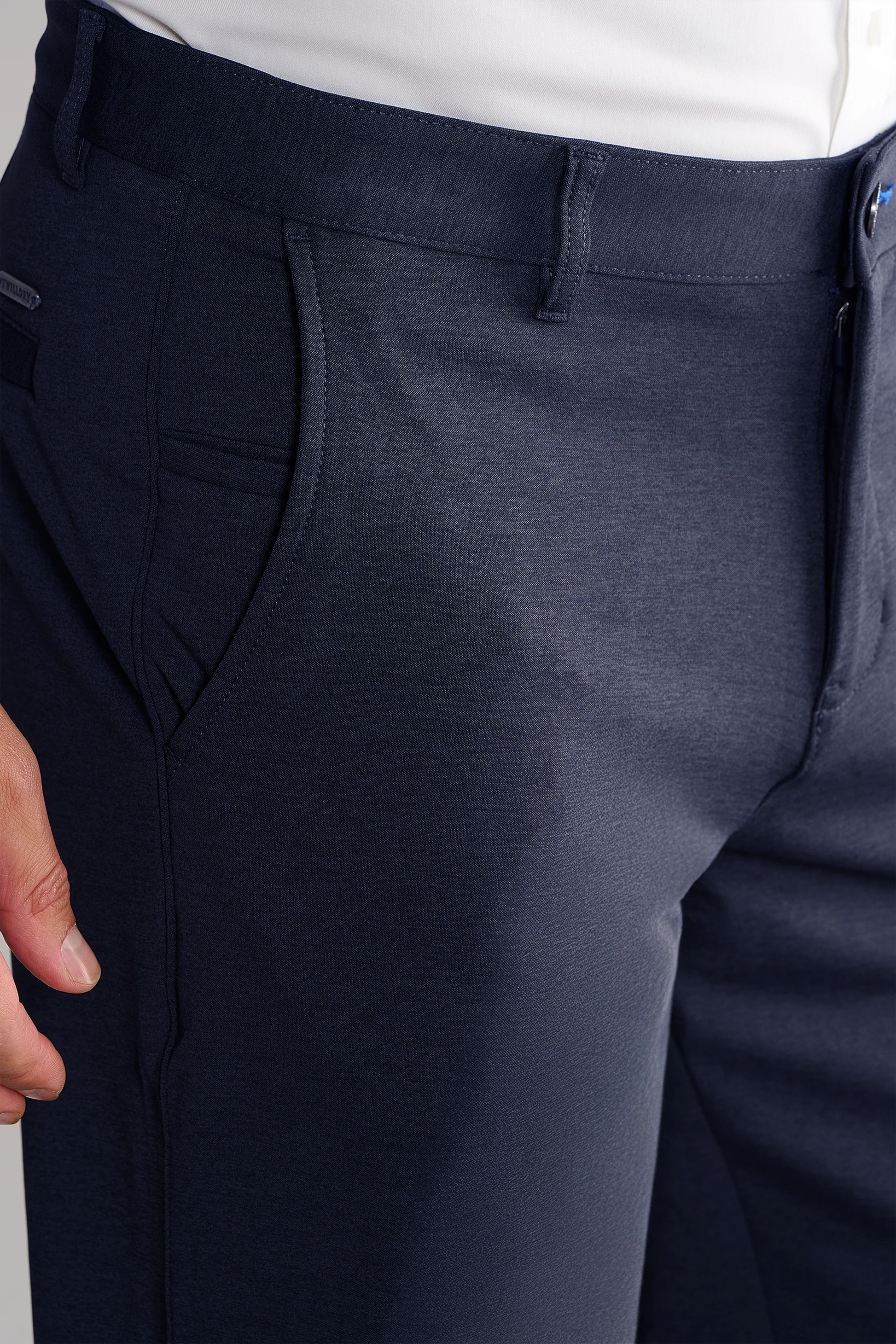 A close-up of a person wearing Twillory Circular Knit Pants in Heather Navy, featuring a tailored fit, wrinkle-resistant fabric with subtle sheen, visible front pocket, and a white tucked-in shirt; their left hand rests near the hip.