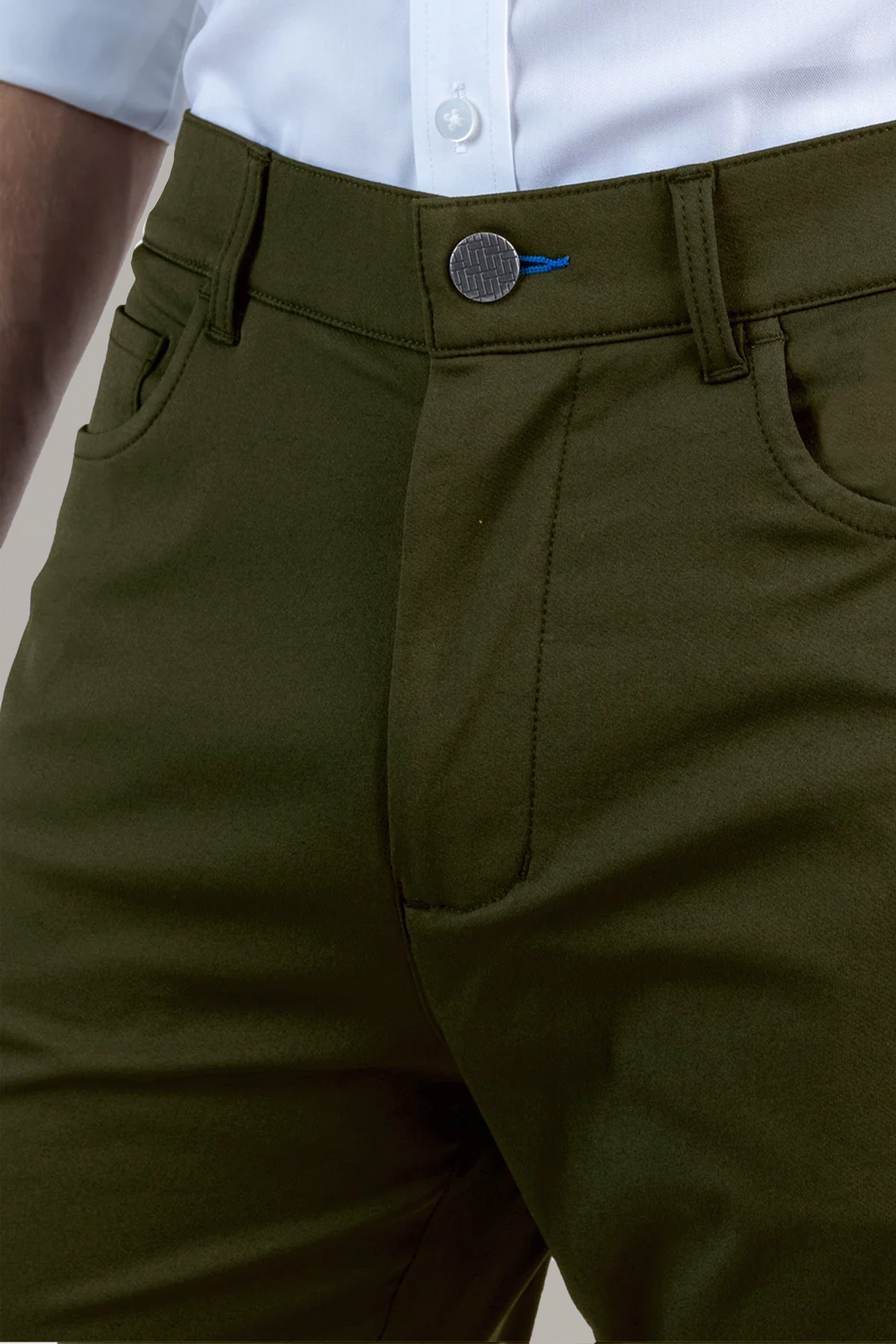 Close-up of a person wearing Twillory Performance Pants // SAGE—olive green, travel-friendly trousers with a round silver button, blue thread detail, belt loops, and front pockets—styled with a tucked-in white shirt.