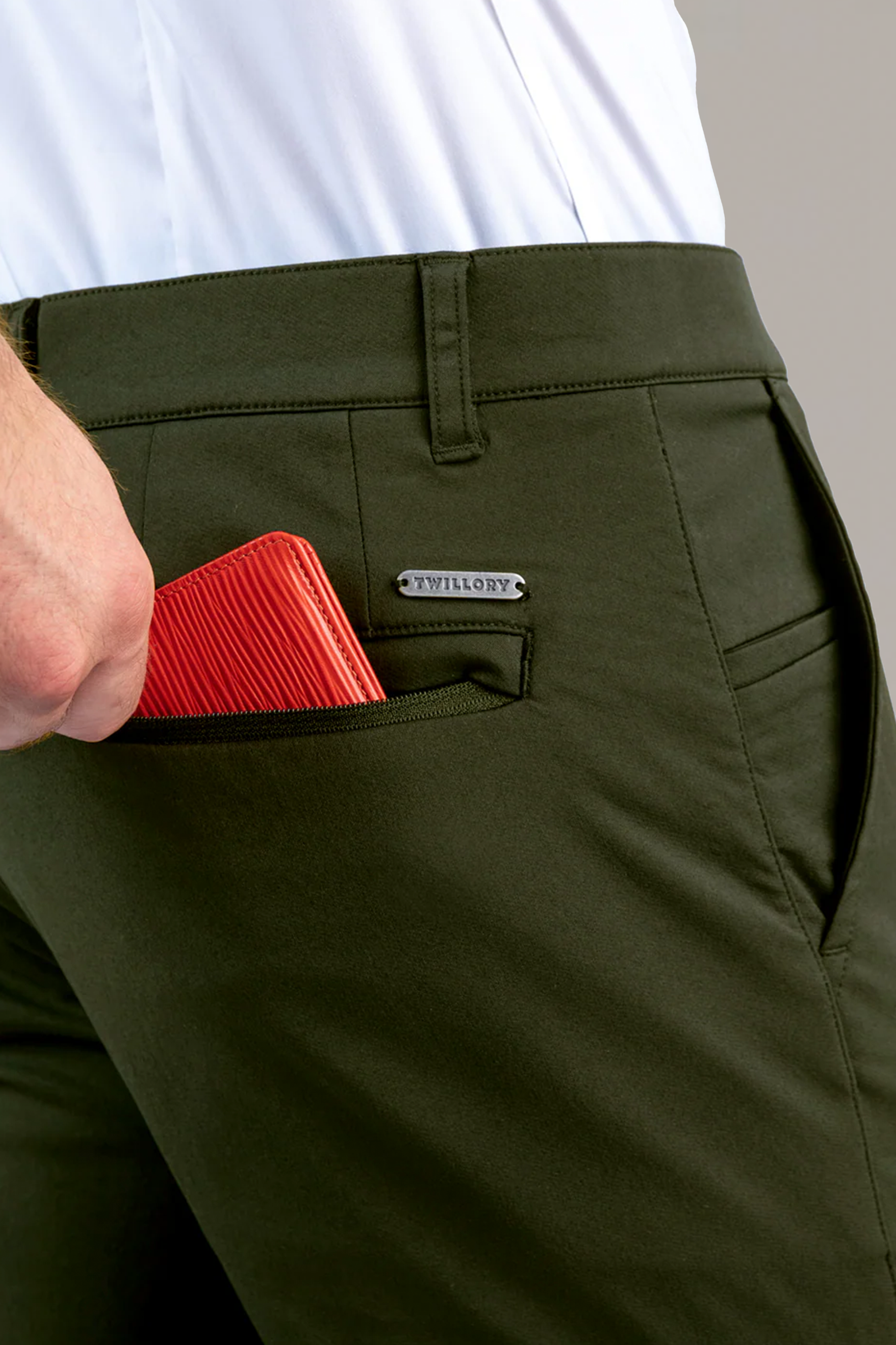 A person wearing Twillory Performance Pants // SAGE and a white shirt is placing a red textured wallet into the front coin pocket, which features a small metal WILHORY tag. Only the lower torso and part of the hand are visible.