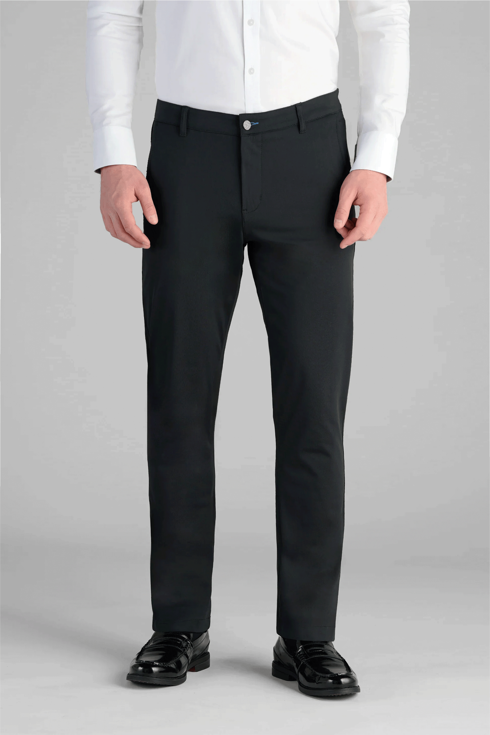 A man stands facing forward, hands at his sides, wearing a white button-up shirt tucked into Twillory Performance Pants in True Black and shiny black dress shoes. The plain light grey background is cropped at the shoulders.