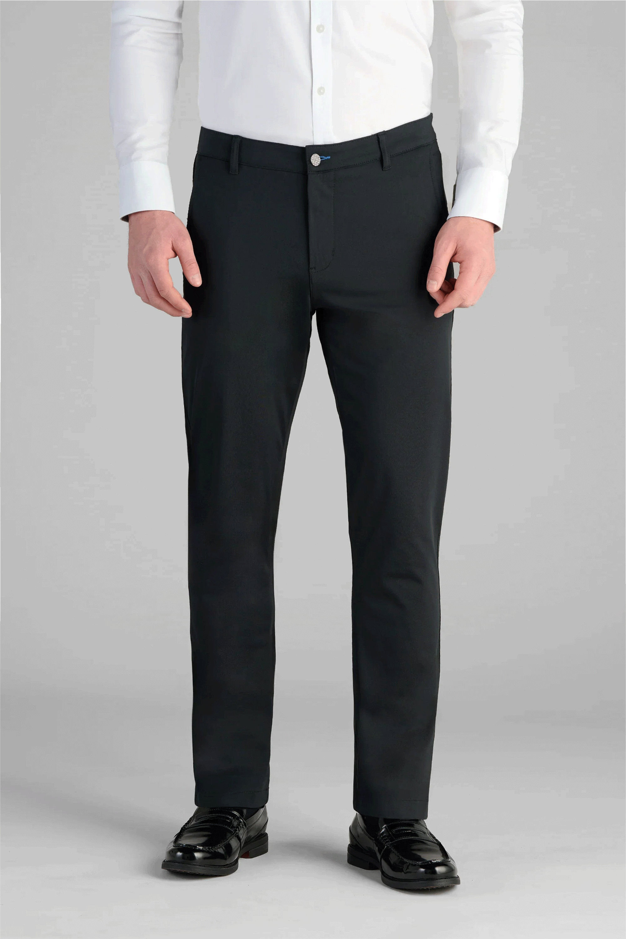 A man stands facing forward, hands at his sides, wearing a white button-up shirt tucked into Twillory Performance Pants in True Black and shiny black dress shoes. The plain light grey background is cropped at the shoulders.