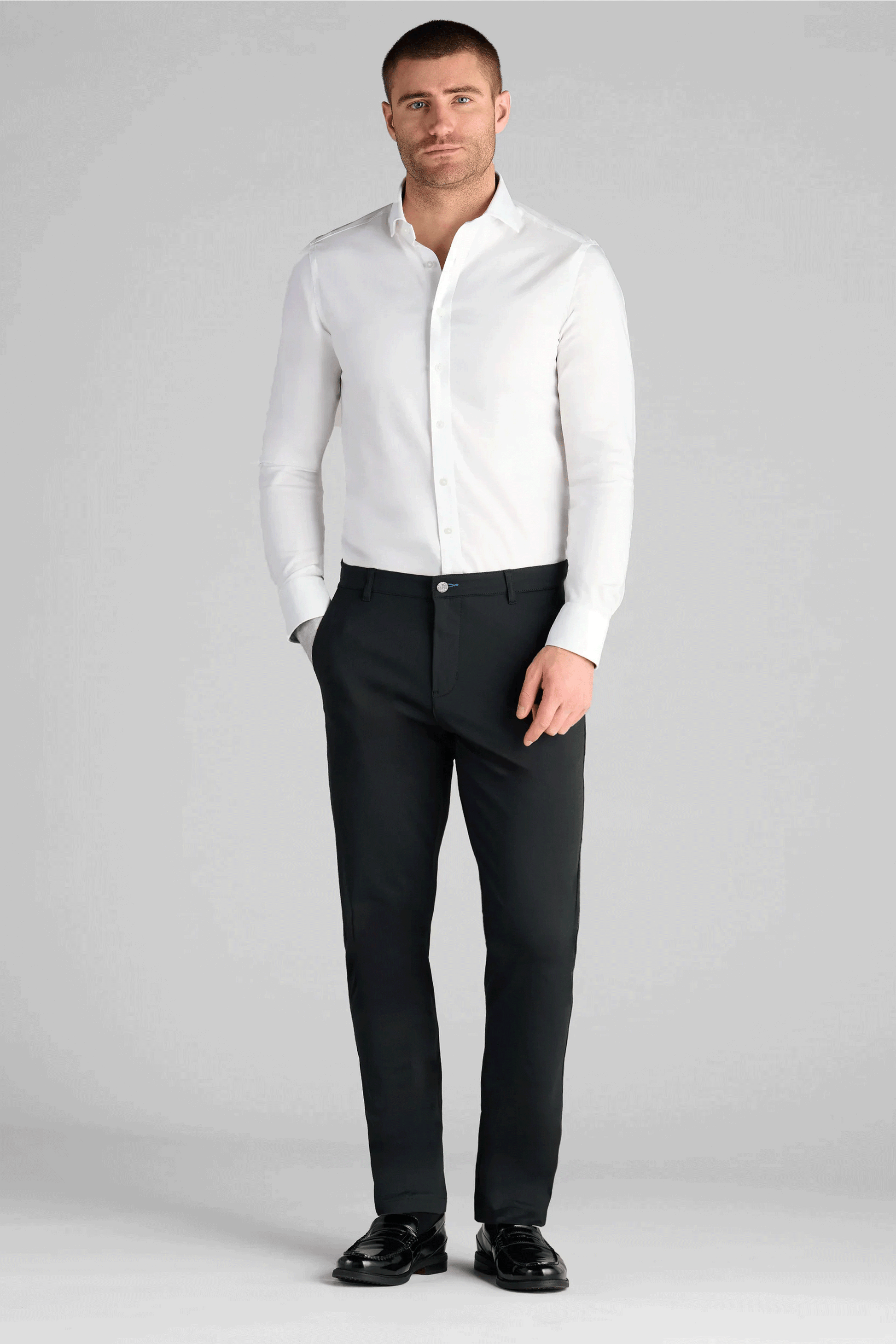 A man stands with one hand in his pocket, wearing a white dress shirt, black Performance Pants // BLACK, and black dress shoes against a plain gray background.
