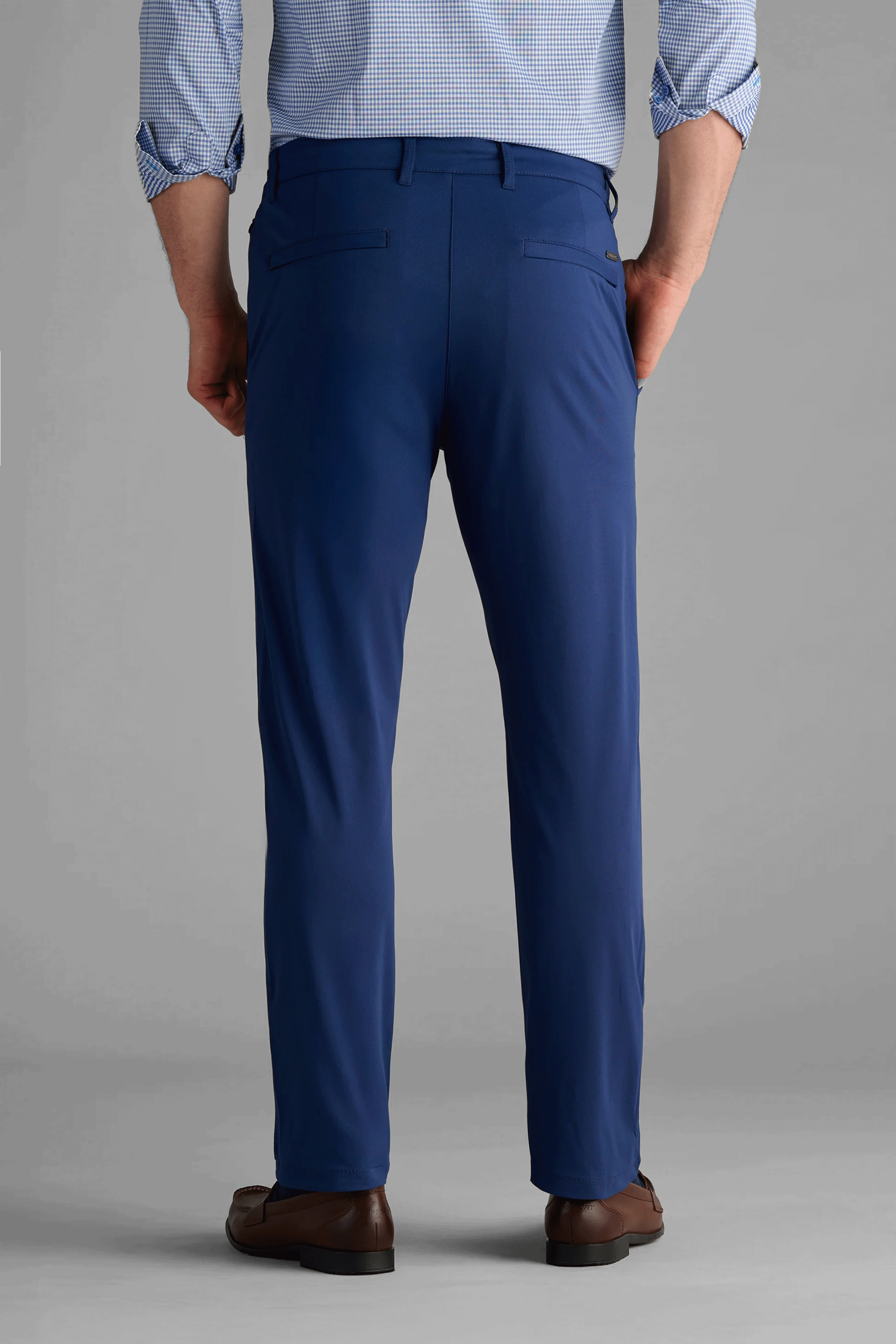 A man faces away, wearing Twillory Performance Pants in cobalt blue and a light blue gingham shirt with rolled sleeves. The trousers feature two back pockets with buttoned flaps. He pairs them with brown dress shoes against a plain light gray background.