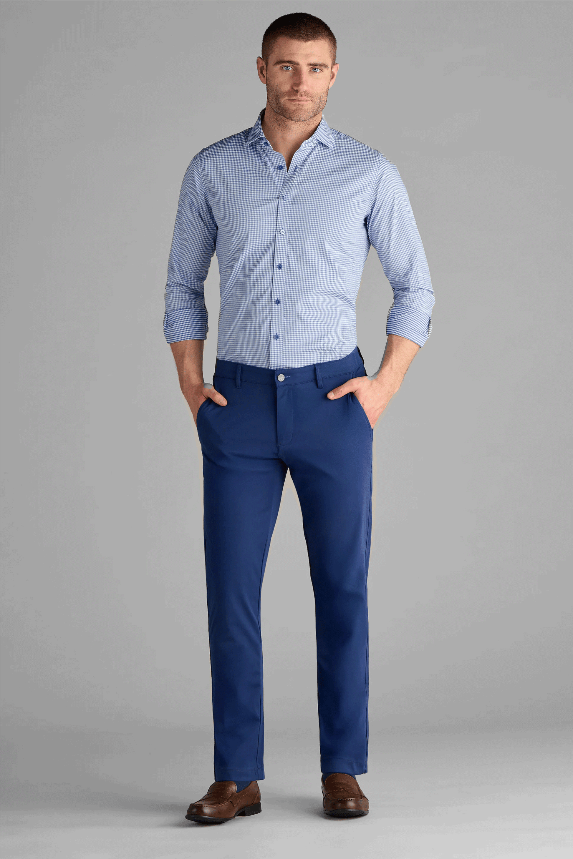 A man with short hair and a trimmed beard poses with hands in pockets, wearing Twillory’s Performance Pants in Cobalt Blue, a light blue button-up shirt with rolled sleeves, and brown dress shoes against a plain gray background.