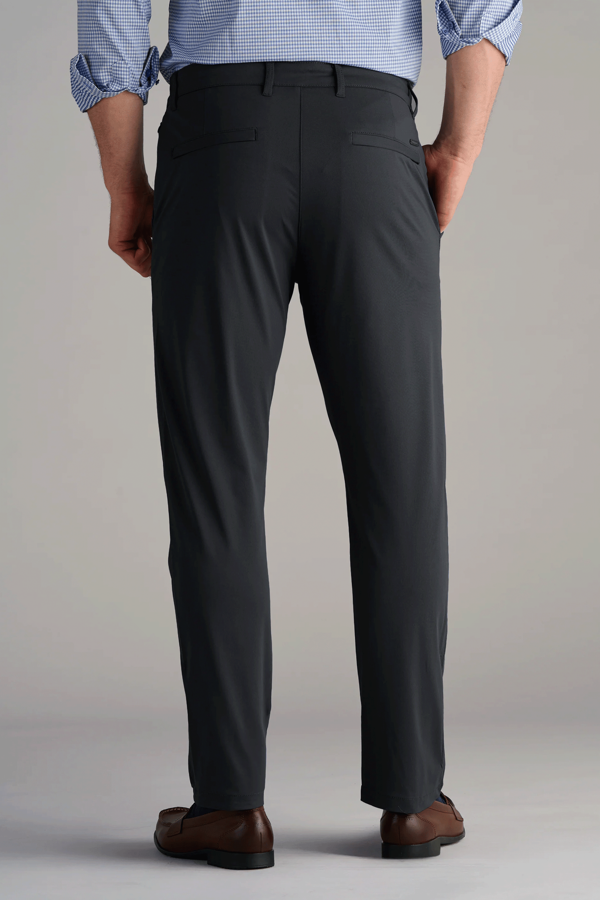 A man stands with his back to the camera, wearing Twillory Performance Pants // Charcoal—dark gray, 4-way stretch dress pants with two buttonless back pockets—paired with a light blue checkered shirt and brown dress shoes against a plain background.