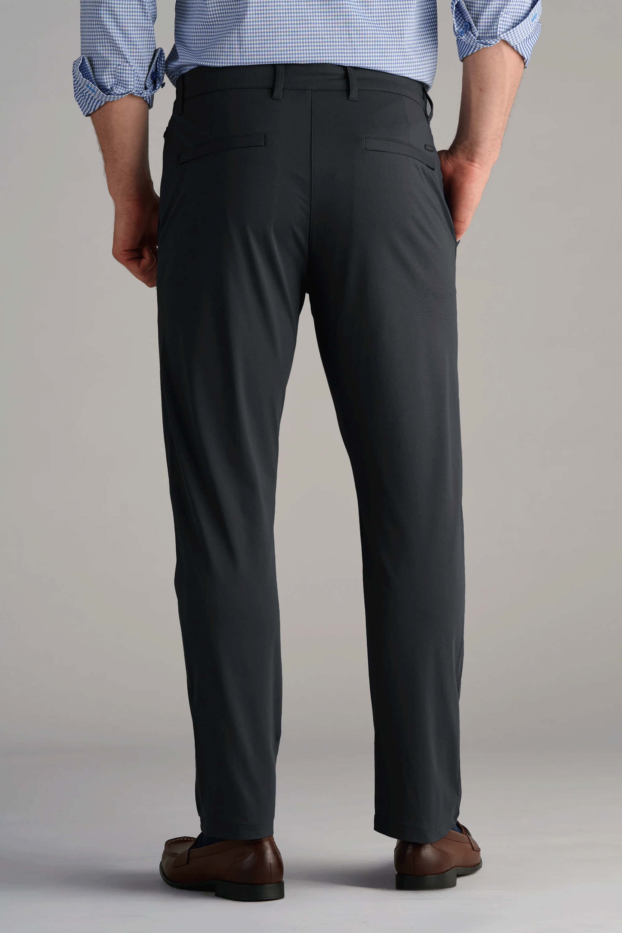 A man stands with his back to the camera, wearing Twillory Performance Pants // Charcoal—dark gray, 4-way stretch dress pants with two buttonless back pockets—paired with a light blue checkered shirt and brown dress shoes against a plain background.