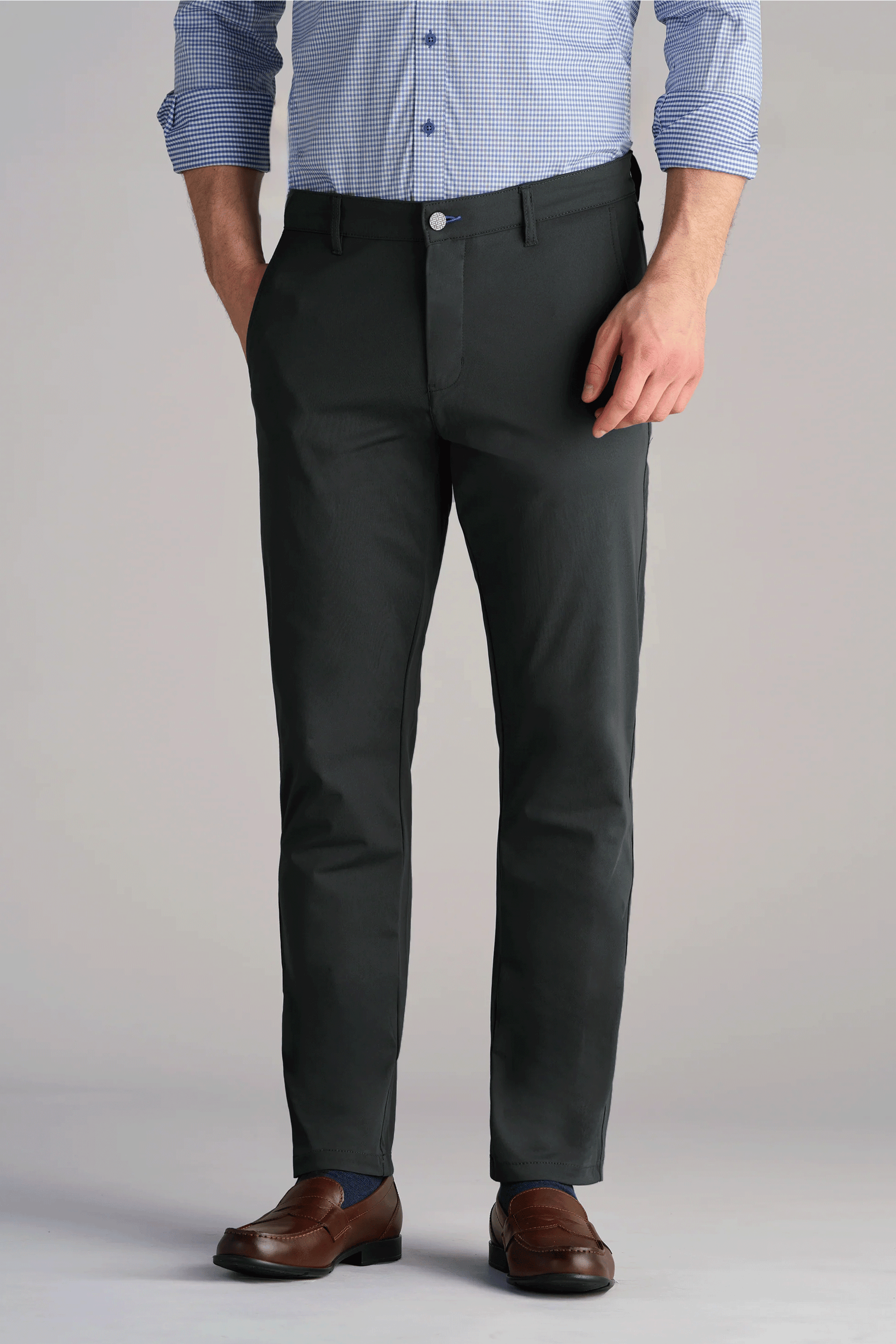 A person wears Twillory Performance Pants // Charcoal—dark gray, slim-fit, 4-way stretch trousers. Paired with a blue-white check shirt (sleeves rolled), brown leather dress shoes. Hands in pockets; shown from mid-chest down.