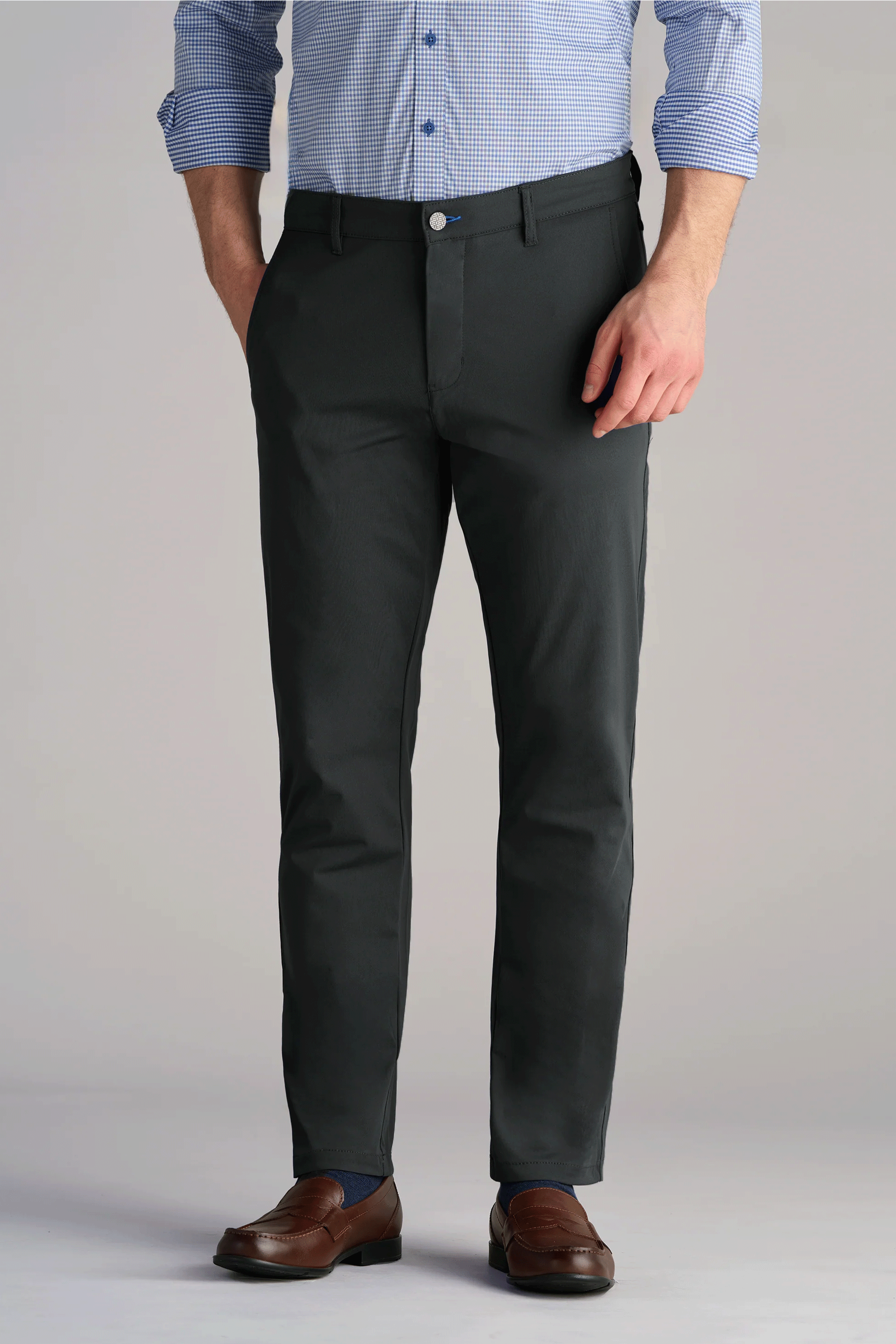 A person wears Twillory Performance Pants // Charcoal—dark gray, slim-fit, 4-way stretch trousers. Paired with a blue-white check shirt (sleeves rolled), brown leather dress shoes. Hands in pockets; shown from mid-chest down.