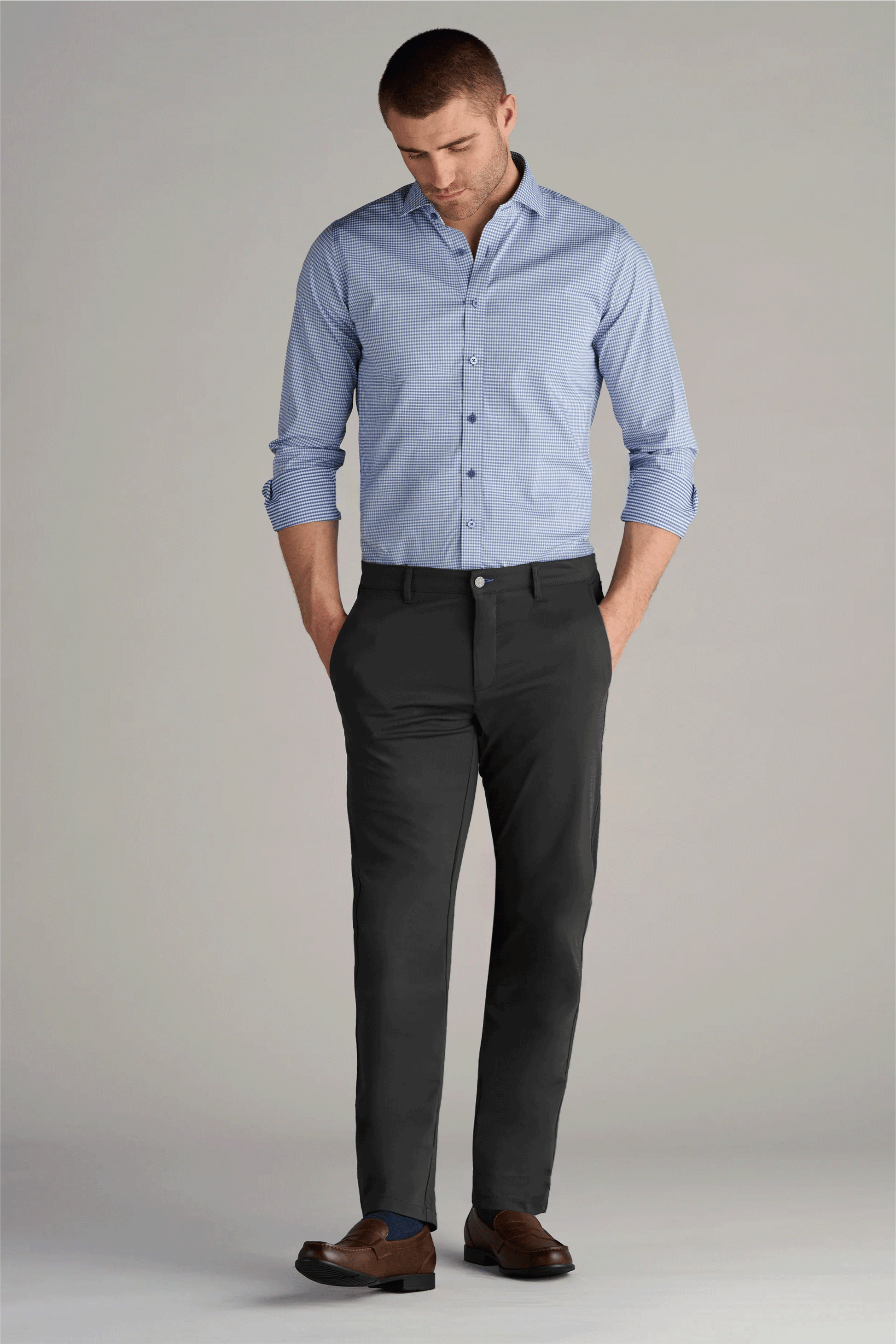 A man stands with his hands in his pockets, wearing a light blue button-up shirt, Twillory Performance Pants in Charcoal with 4-way stretch, and brown loafers. He looks down slightly against a plain light gray background.