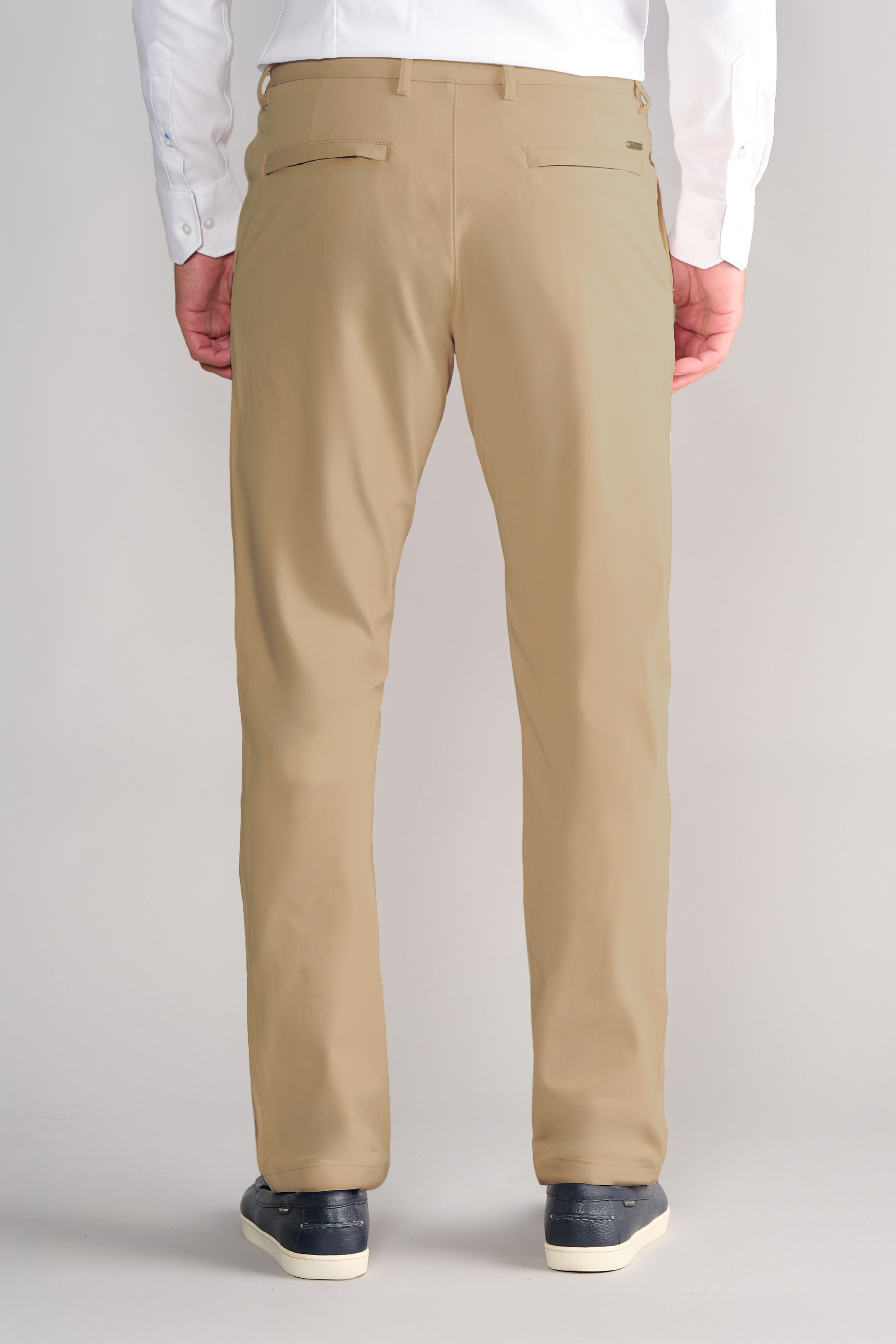 A person faces away, wearing Twillory Performance Pants in Khaki with two back welt pockets, a white button-up shirt, and dark shoes with white soles. Hands rest at sides; the background is plain and light-colored.