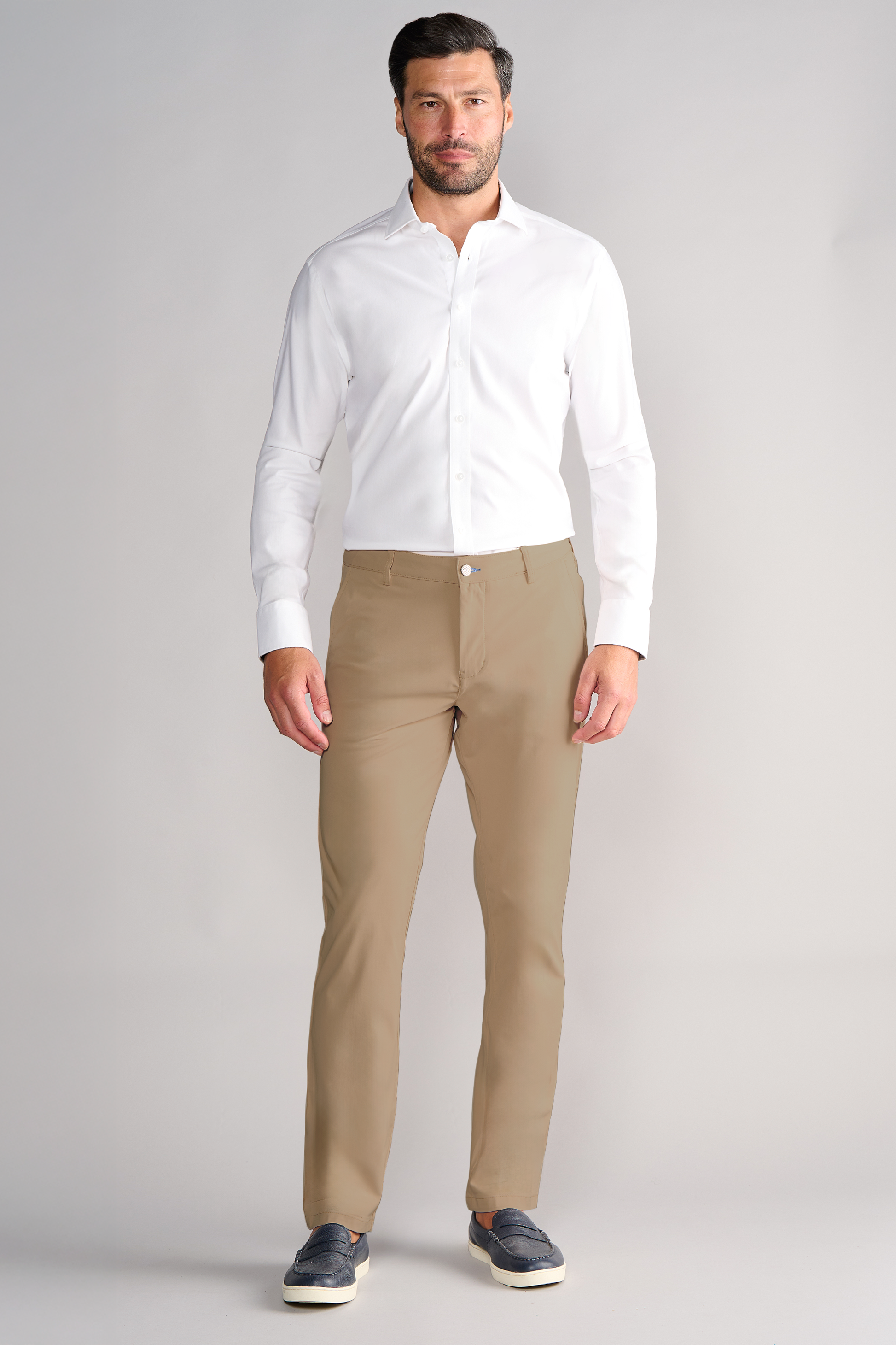 A man with a neat beard and short hair faces forward, wearing a fitted white shirt, Twillory Performance Pants in Khaki, and dark slip-on shoes. His sleeves are down, standing against a plain light gray studio portrait background.