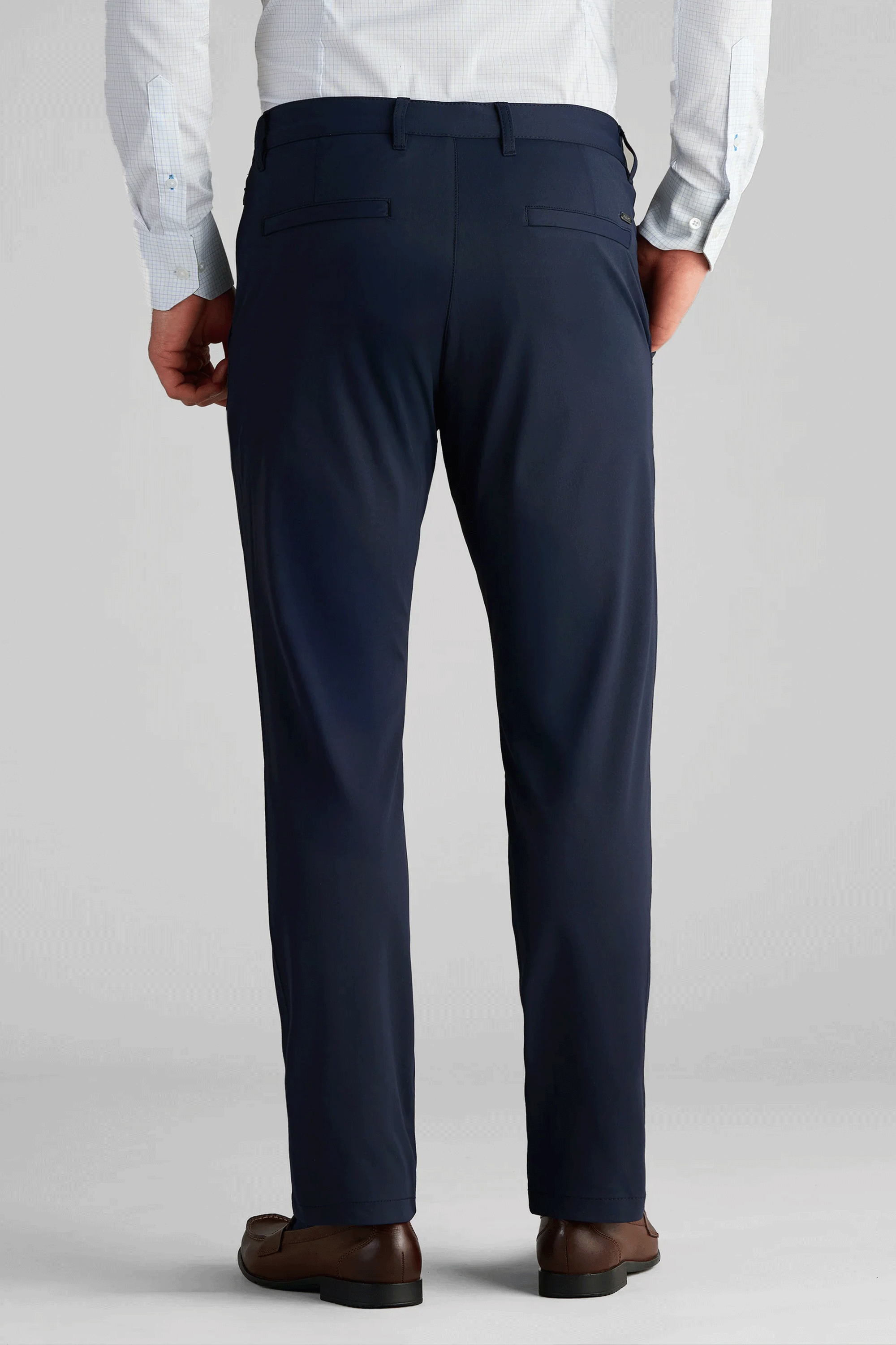 A man, facing away, wears Twillory Performance Pants in navy blue with two back welt pockets and belt loops, paired with a light blue patterned button-up and brown dress shoes. The background is plain light gray.