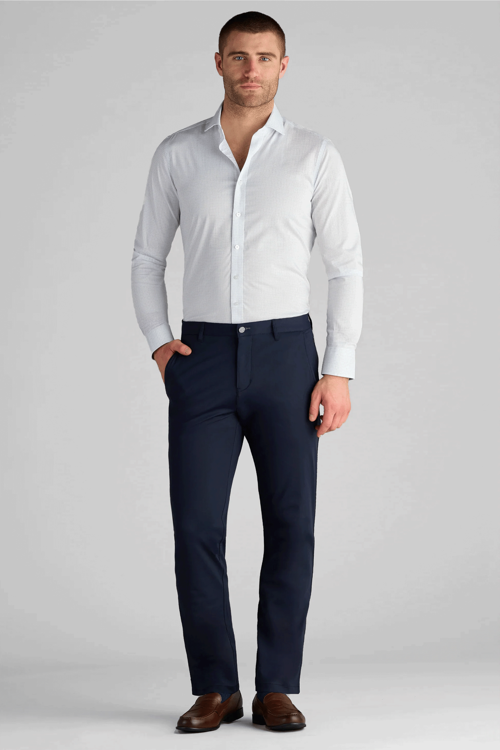 A man with short brown hair and a trimmed beard stands facing forward, hands in pockets, wearing Twillory Performance Pants in navy blue with a light grey dress shirt tucked in and brown dress shoes. The background is plain light grey.