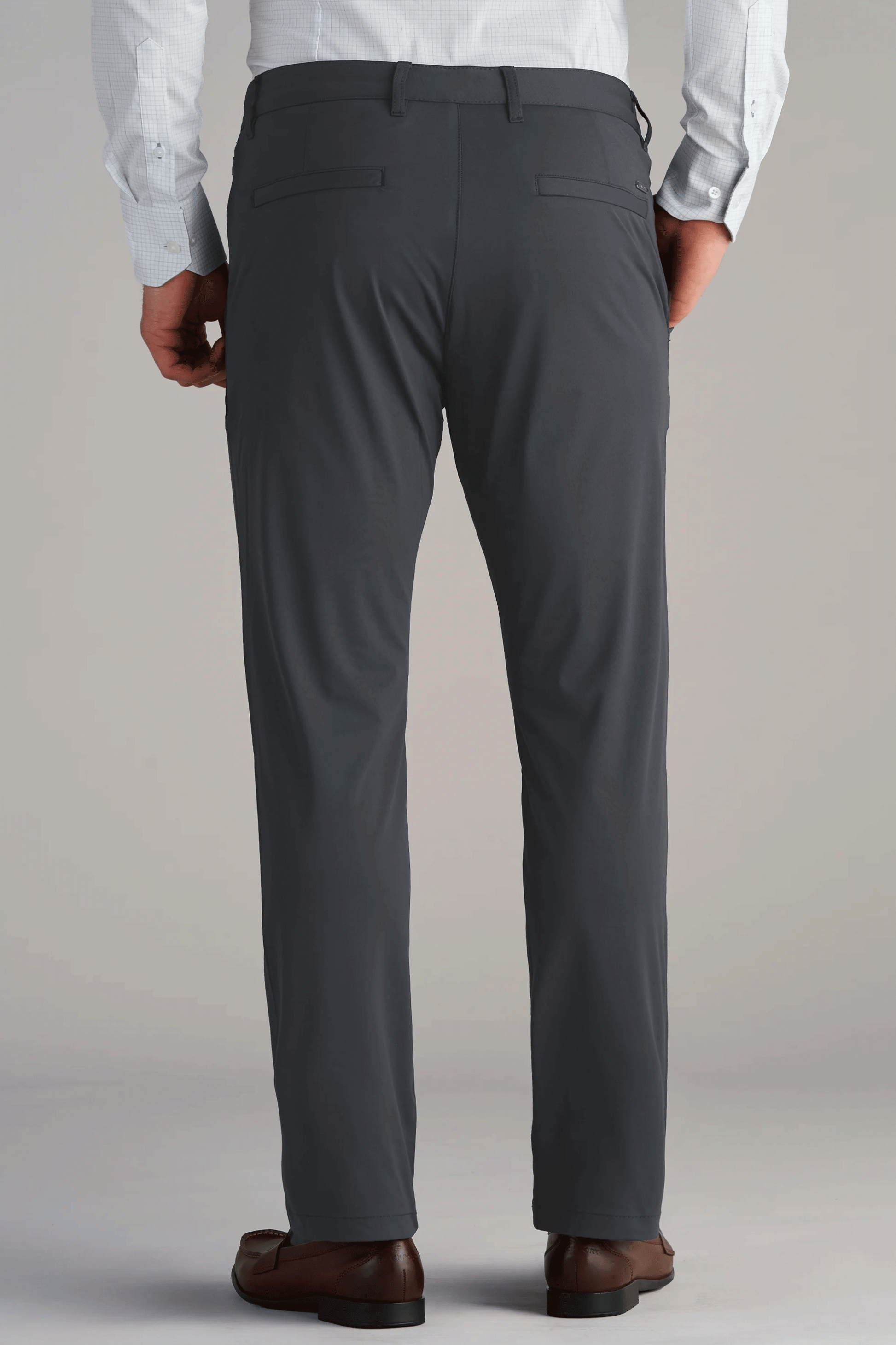 A person stands with hands at their sides, wearing Twillory Performance Pants in SLATE GREY, a light blue tucked-in shirt, and brown dress shoes. The simple light background highlights the stylish pants.