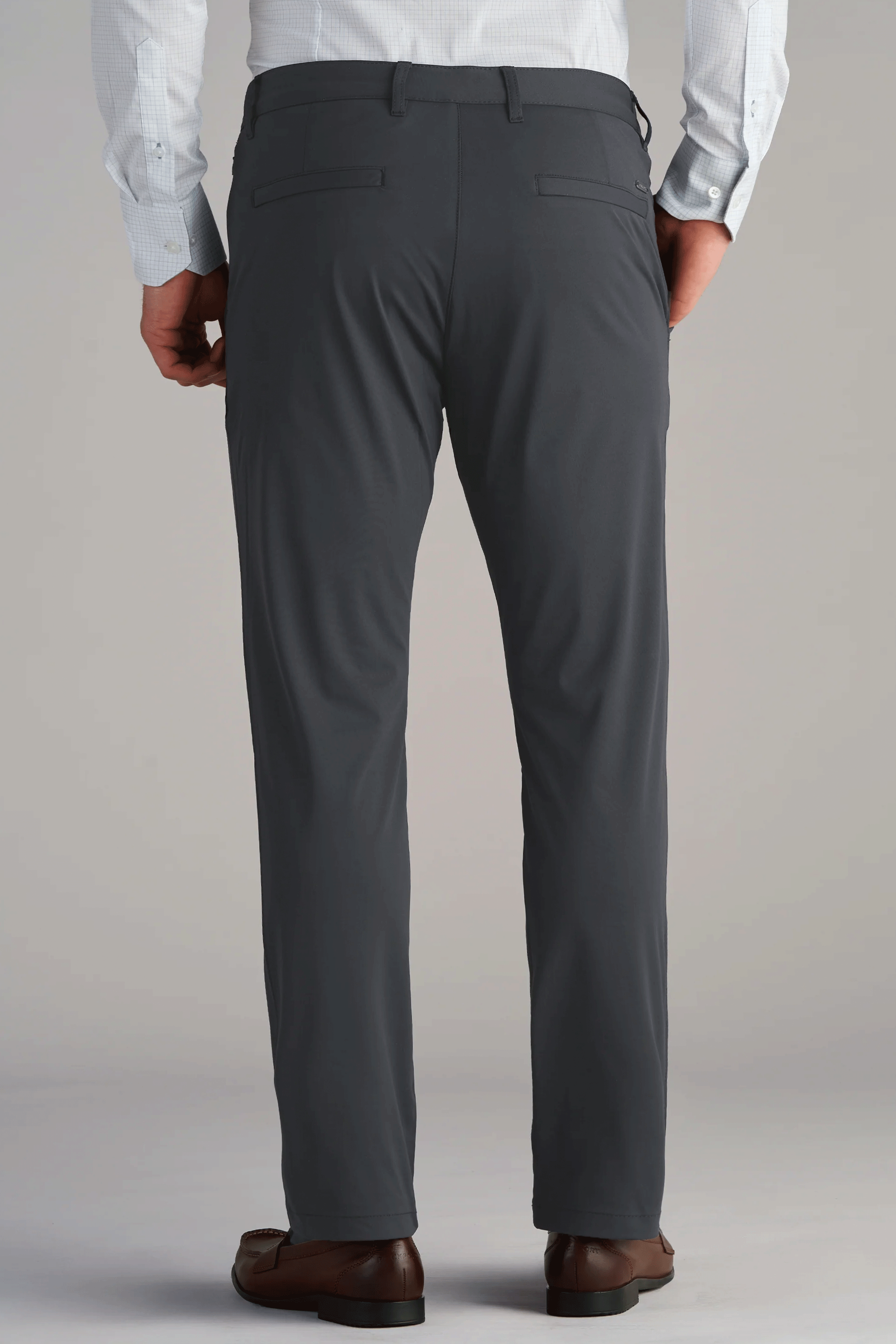 A person stands with hands at their sides, wearing Twillory Performance Pants in SLATE GREY, a light blue tucked-in shirt, and brown dress shoes. The simple light background highlights the stylish pants.