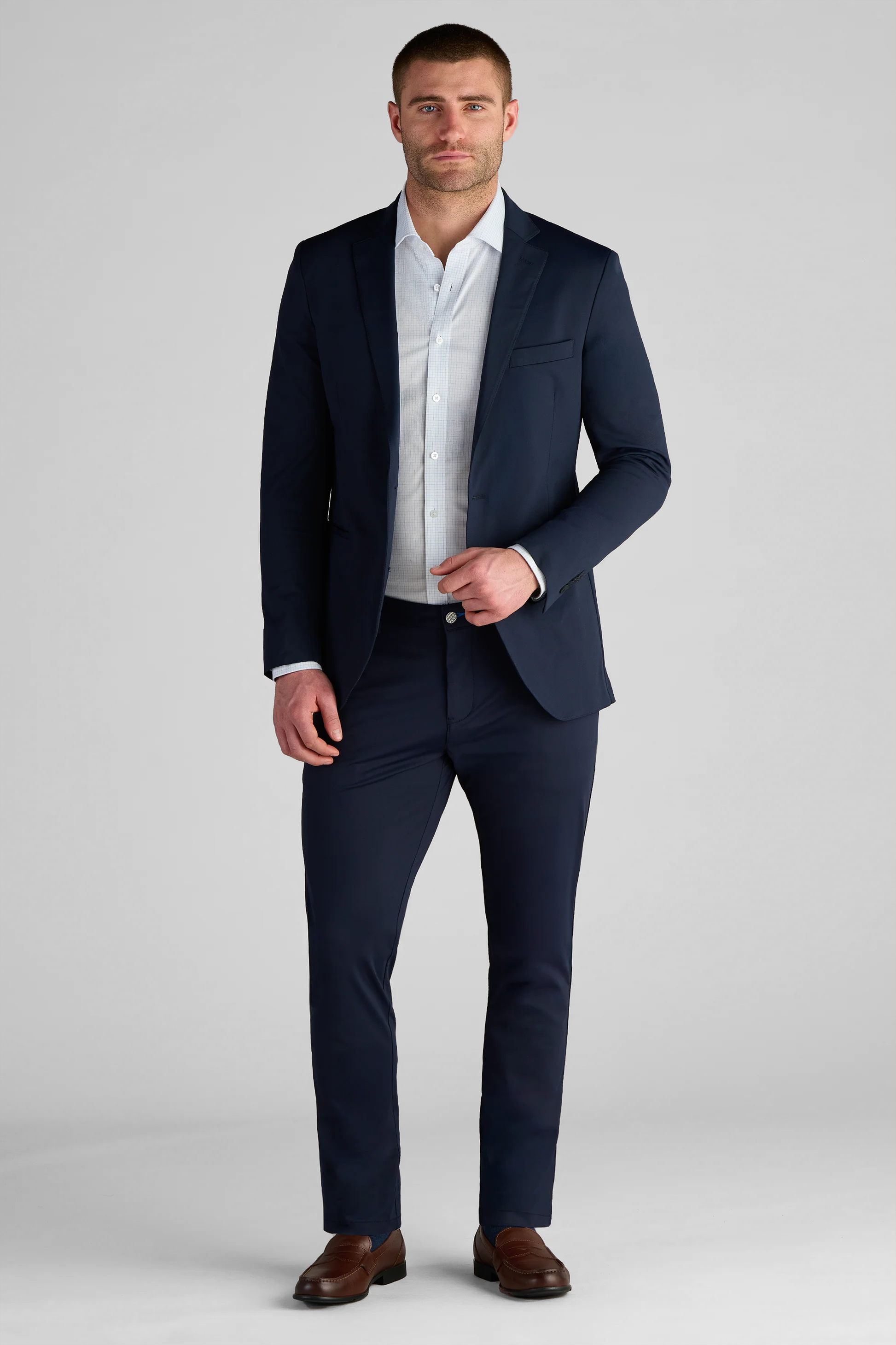 A man with short brown hair and a trimmed beard stands facing forward in a Twillory Performance Blazer // Navy Blue over a white dress shirt, no tie. He holds the jacket with his left hand, wears brown loafers, and stands against a light gray background.