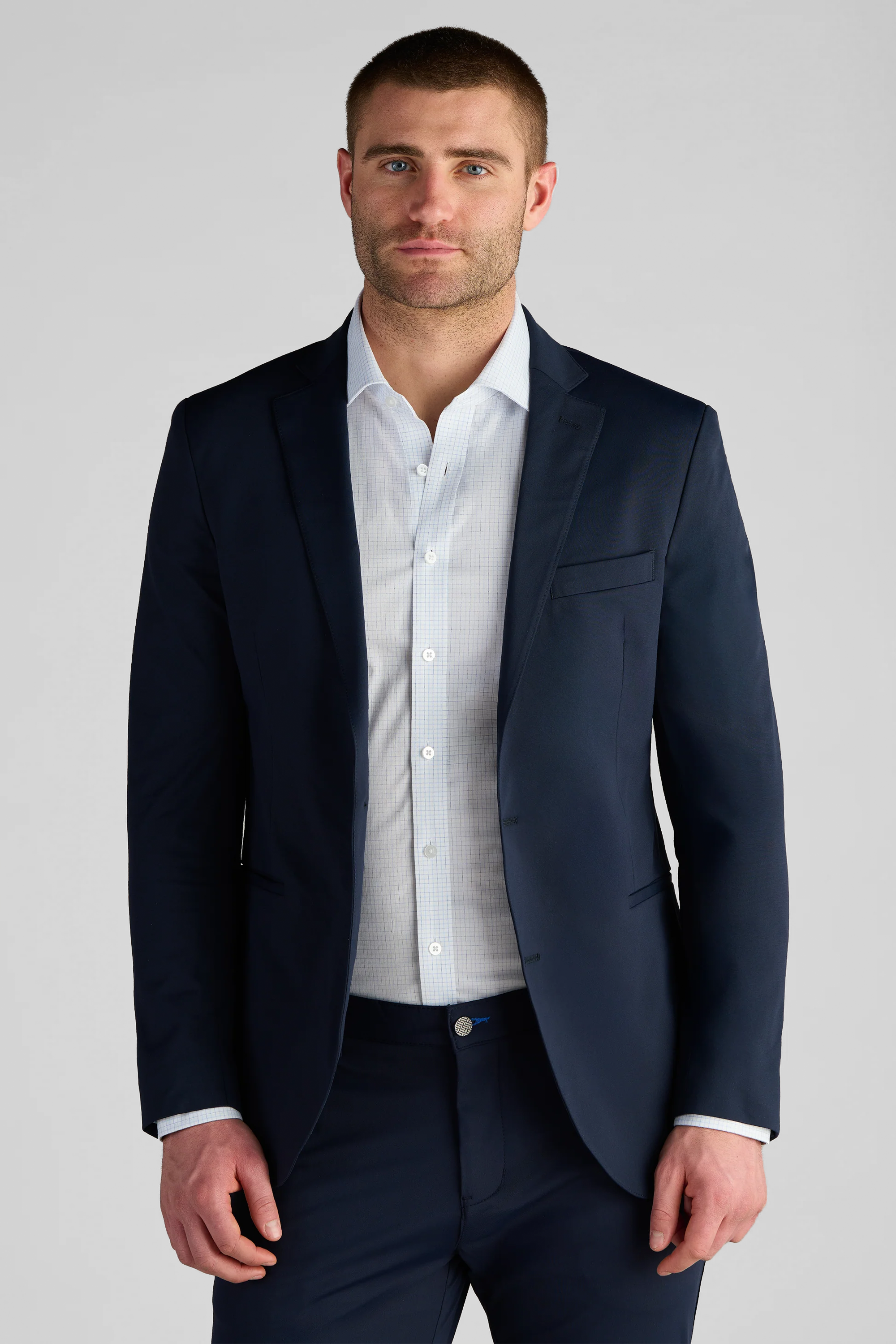 A man with short hair and a beard faces forward in a Twillory Performance Blazer // Navy Blue over a light blue button-up. The stretch fabric blazer is cuffed, untied at the neck, set against a plain light gray background.