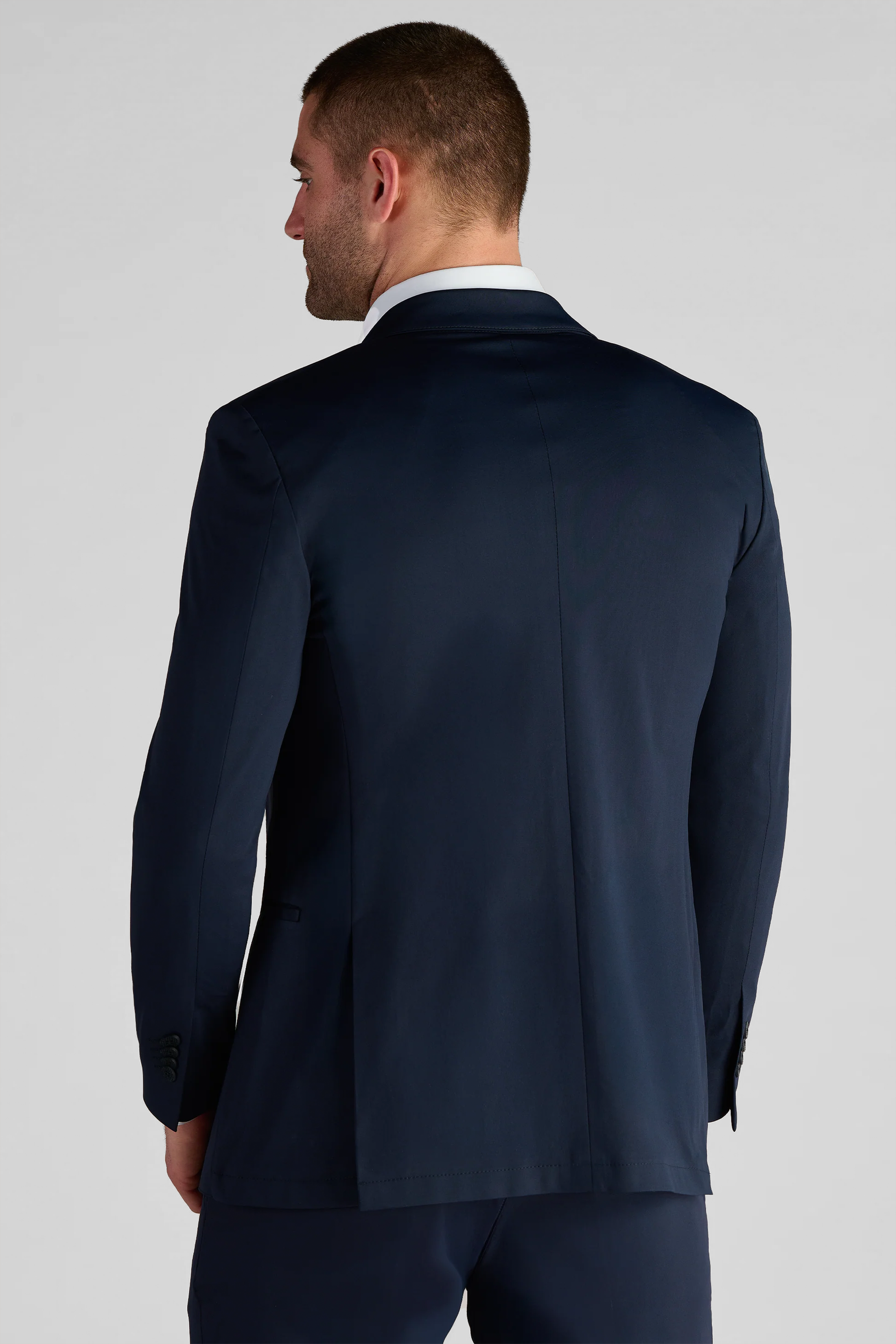 A man with short, neat hair faces away from the camera, wearing the Twillory Performance Blazer // Navy Blue and matching trousers. The blazer features a single back vent and buttoned sleeve cuffs. The background is plain and light-colored.
