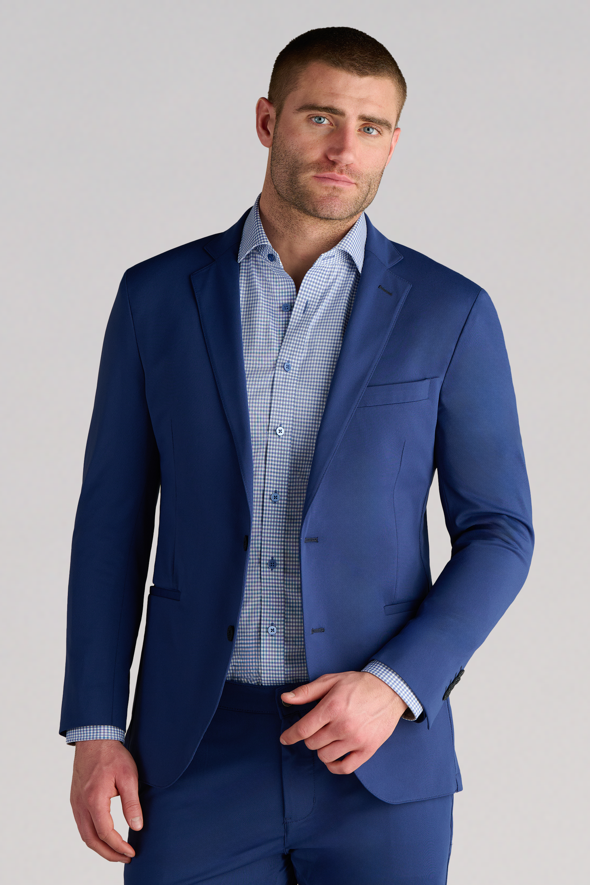 A man with short brown hair and light stubble wears the Twillory Performance Blazer in Cobalt Blue, paired with matching trousers and a light blue checkered shirt. He stands against a plain gray background, looking forward with a slight smile.