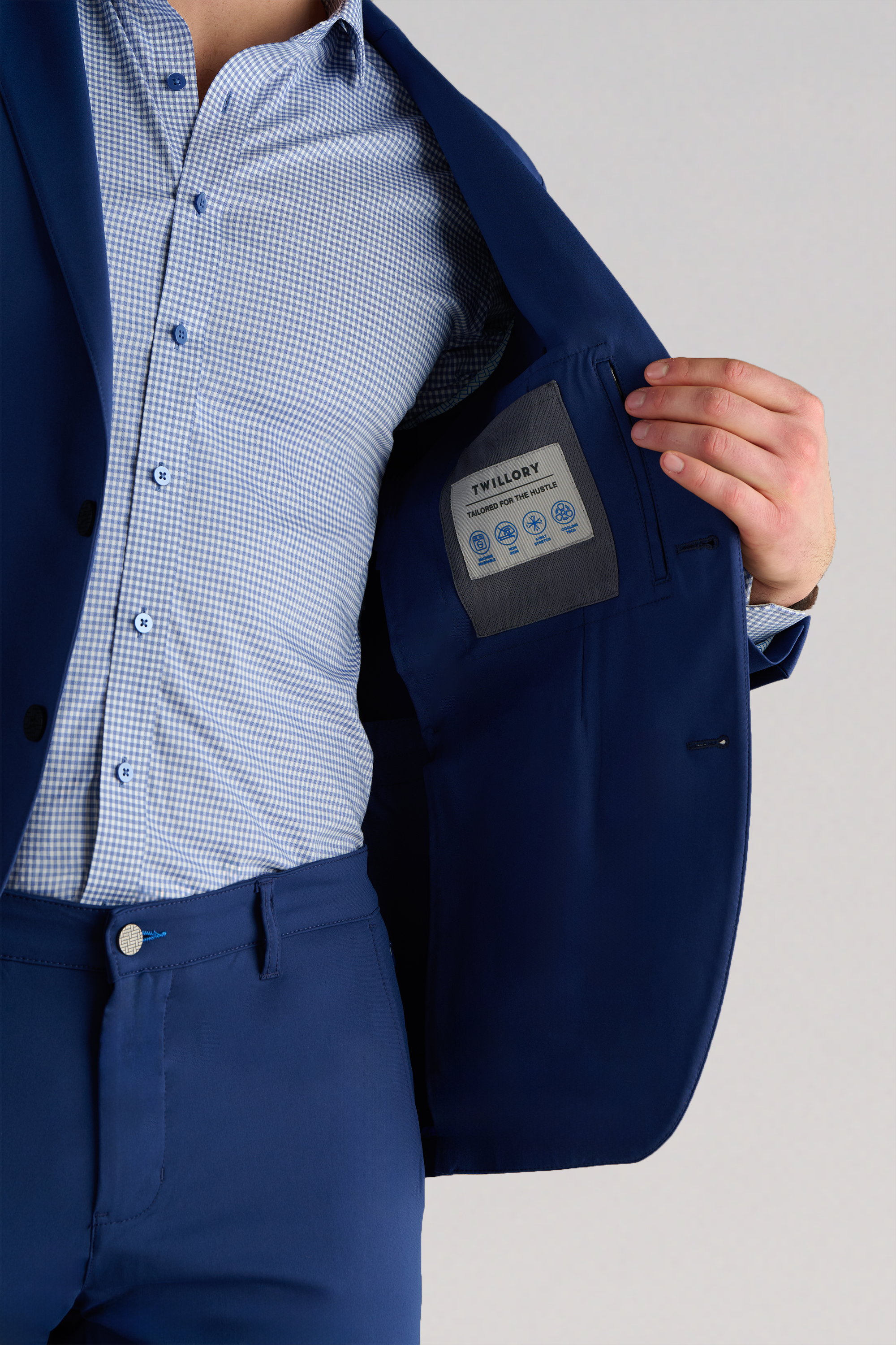 A person wearing the Twillory Performance Blazer in Cobalt Blue and a light blue checkered shirt holds open the jacket to show the inner pocket with a sewn-in label. The background is plain and light-colored.