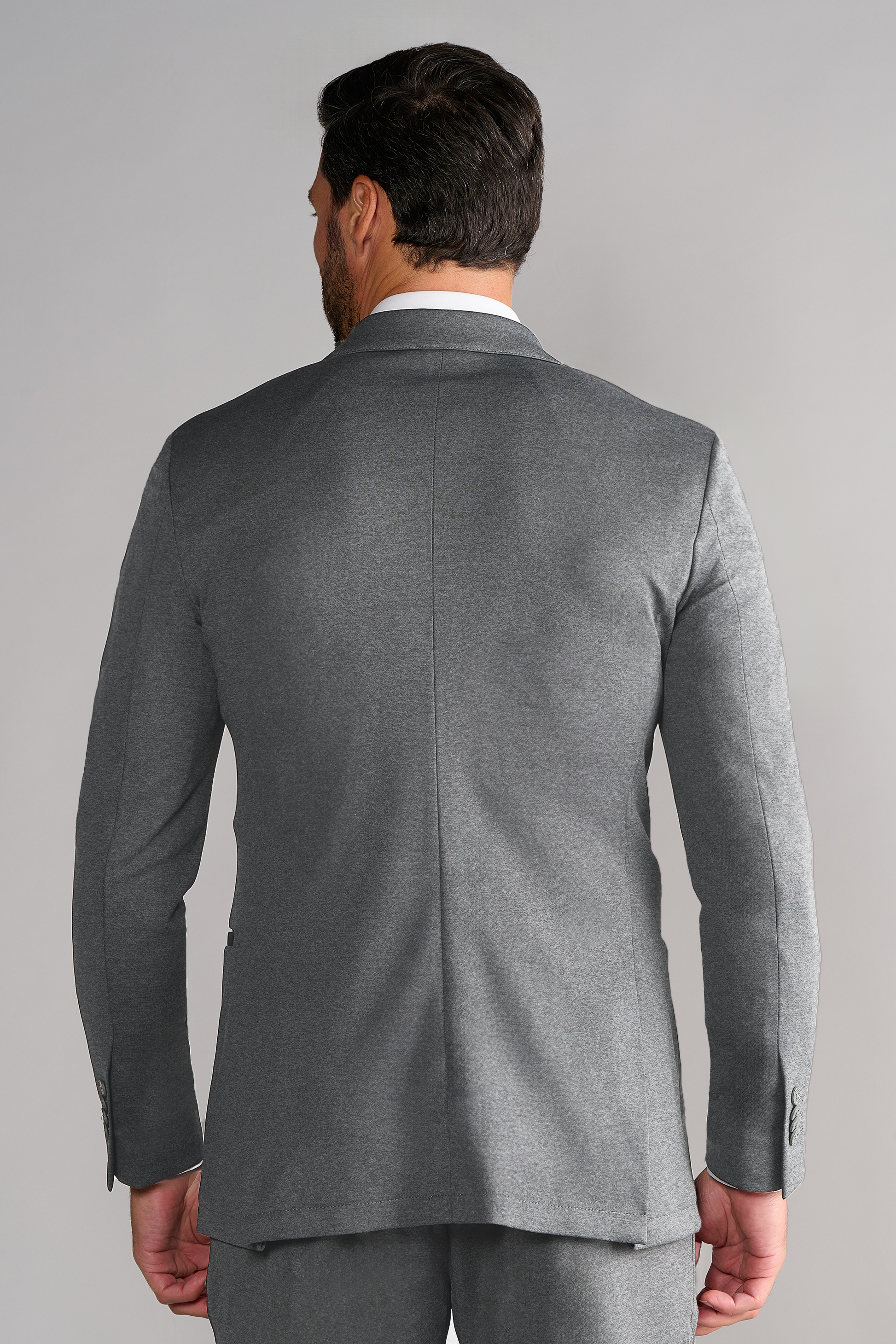 A man with dark hair and a beard is seen from behind wearing Twillory’s Performance Blazer // Heather Light Grey. The plain backdrop highlights the suit’s center back seam and modern stretch design.