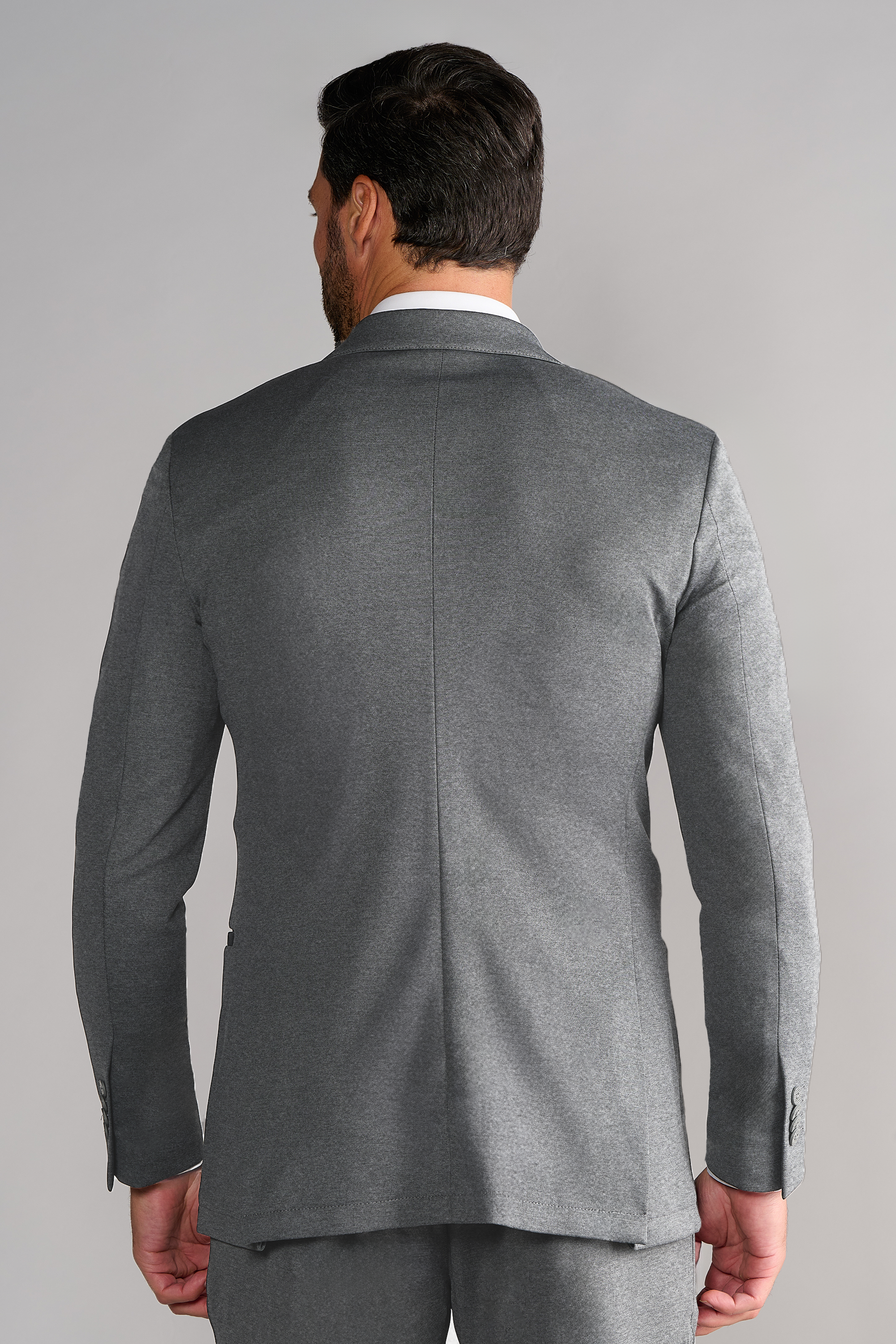 A man with dark hair and a beard is seen from behind wearing Twillory’s Performance Blazer // Heather Light Grey. The plain backdrop highlights the suit’s center back seam and modern stretch design.
