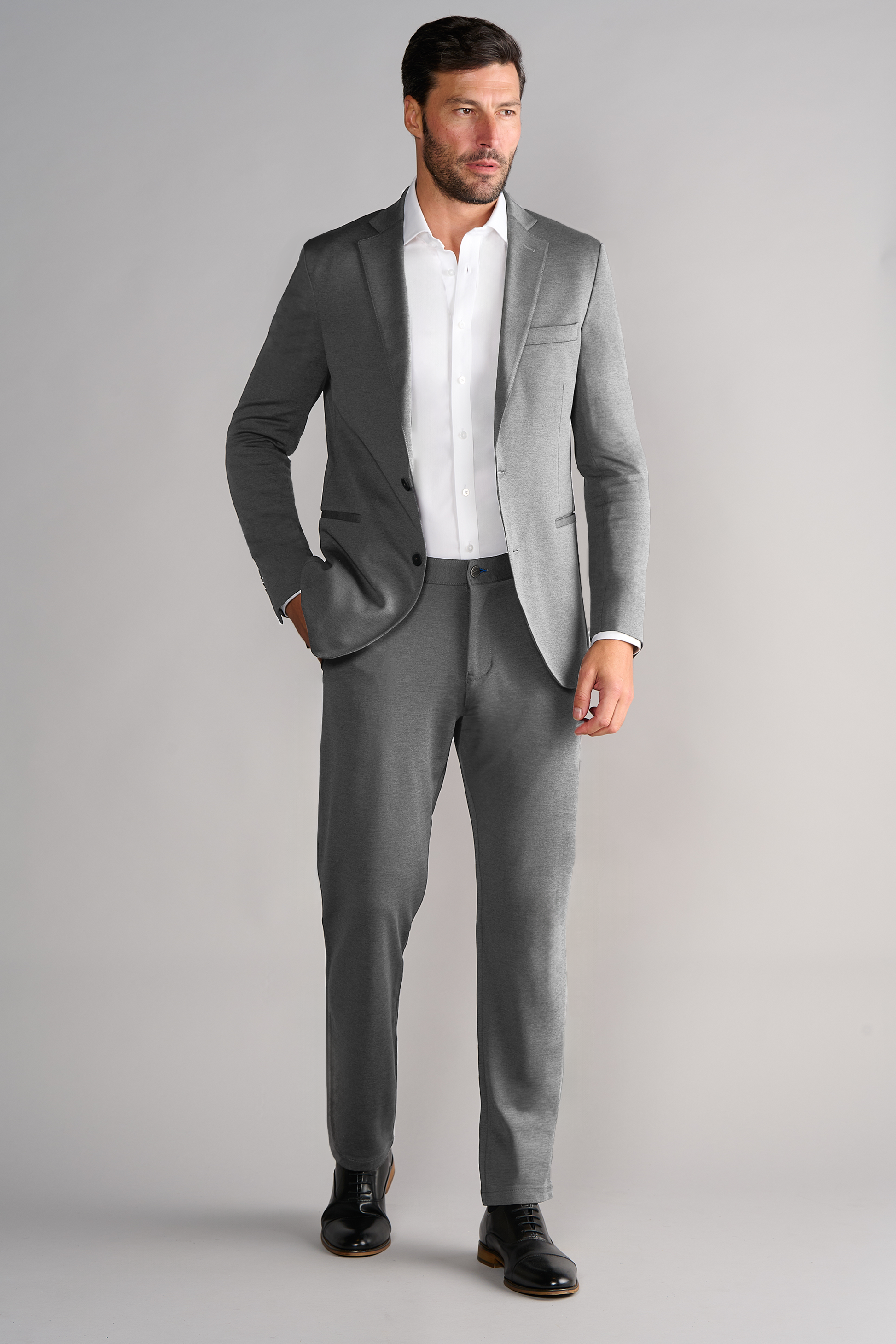 A man with dark hair and a trimmed beard models the Twillory Performance Blazer in Heather Light Grey, paired with a white dress shirt and black shoes. He stands against a light gray backdrop, one hand in his pocket, gazing to the side.